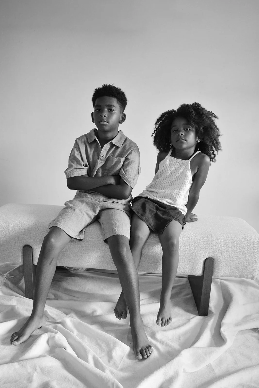 Black and white photo of a young boy and girl sitting on a modern, upholstered bench with wooden legs, posed against a plain background, with a draped cloth on the floor underneath.