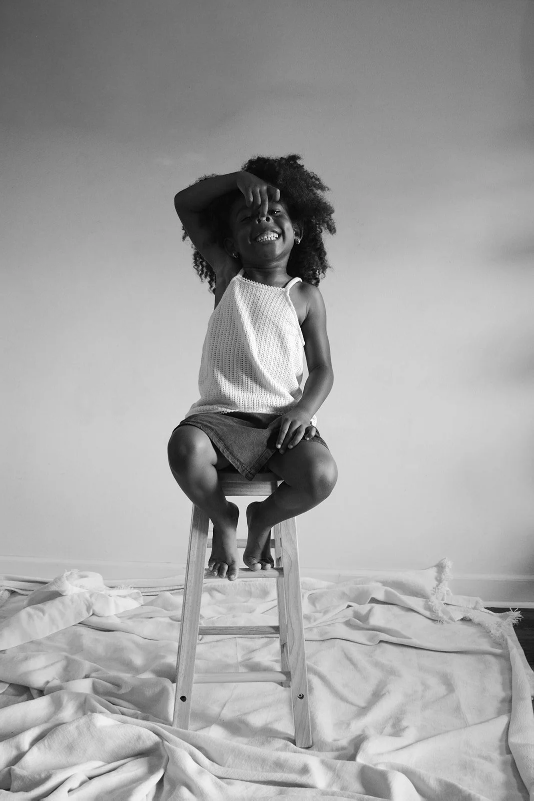 A smiling young girl with curly hair is sitting barefoot on a wooden stool, wearing a sleeveless top and shorts, with cloths spread on the floor underneath, in a plain indoor setting.