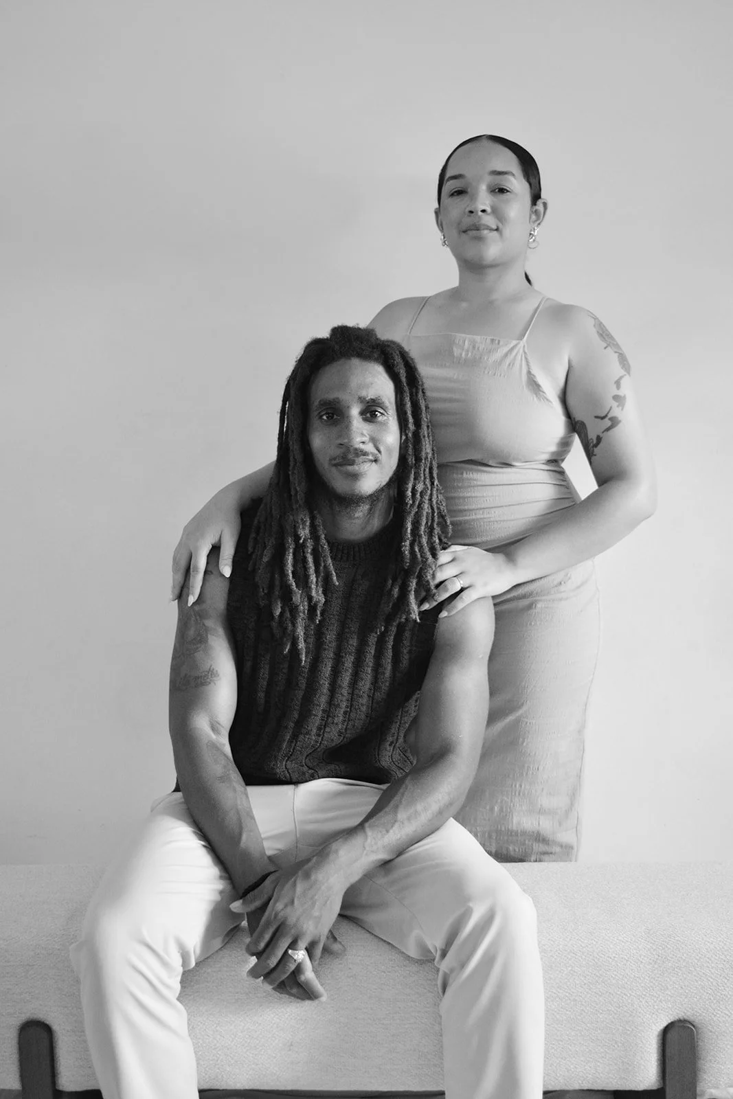 A black and white photo of a man with long dreadlocks sitting on a bench with his hands clasped, and a woman standing behind him with her hand on his shoulder and her other hand on her hip, both looking at the camera.