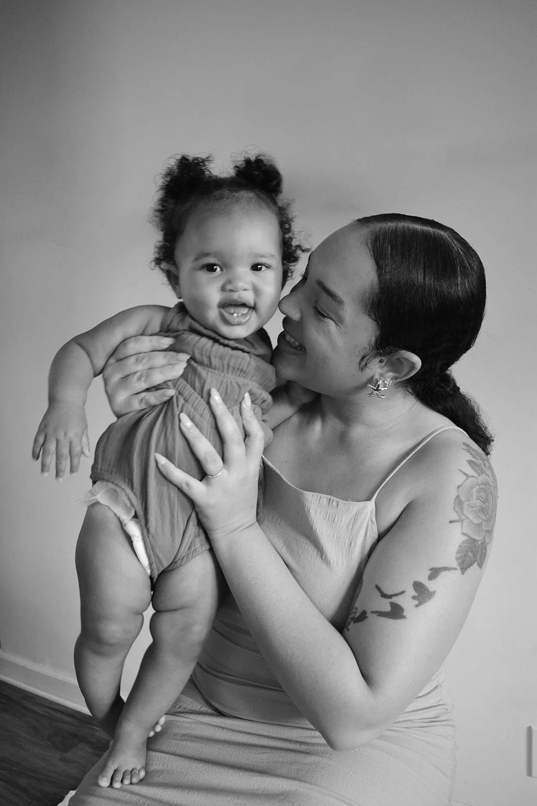 A woman with dark, wavy hair holding up a smiling toddler girl with curly hair in front of a plain background.