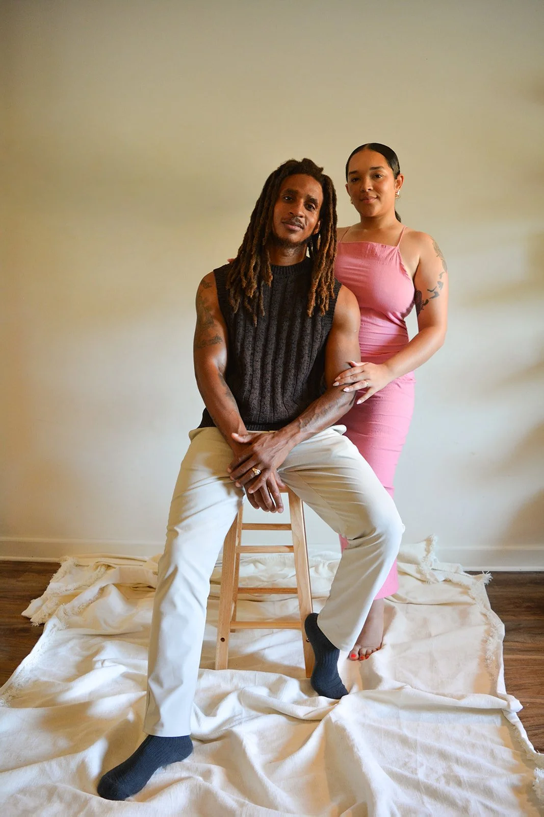 A man with dreadlocks and a black sleeveless shirt sitting on a stool, and a woman in a pink dress standing beside him, holding his arm in an indoor setting with a beige wall and a white cloth on the floor.