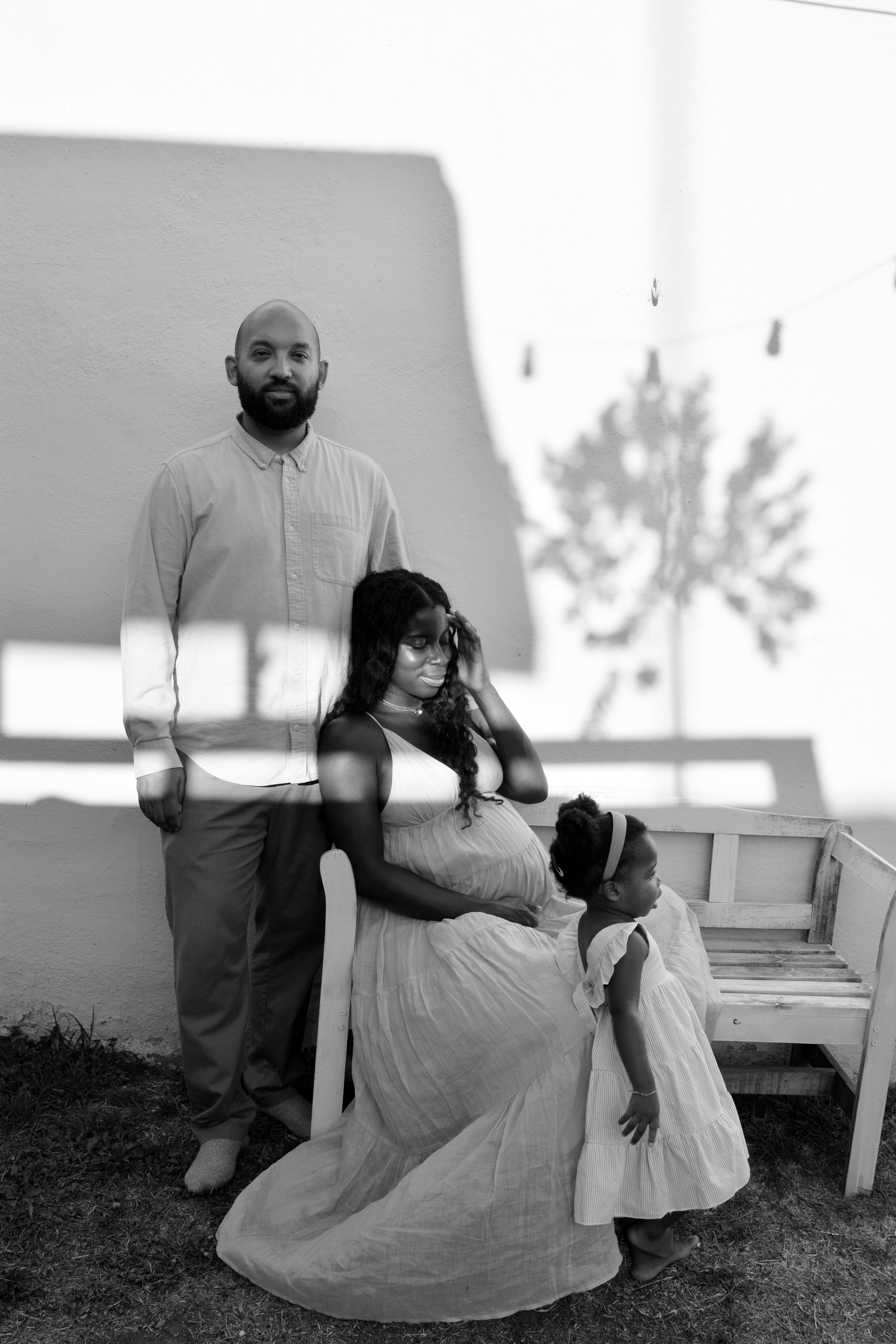 A black-and-white photo of a man, woman, and two children outdoors, with a plain wall and a tree's shadow in the background. The man has a beard and is standing, the woman is sitting on a bench with her head resting on her hand, and the children are 