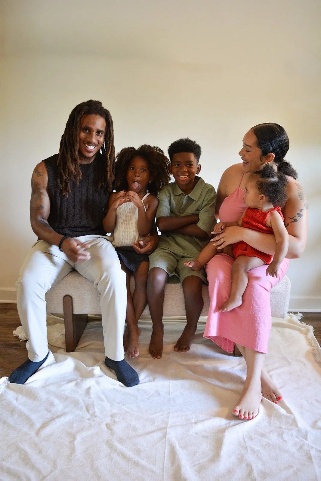 Family of five sitting on a bench, smiling and making funny faces, in a room with a plain beige wall.