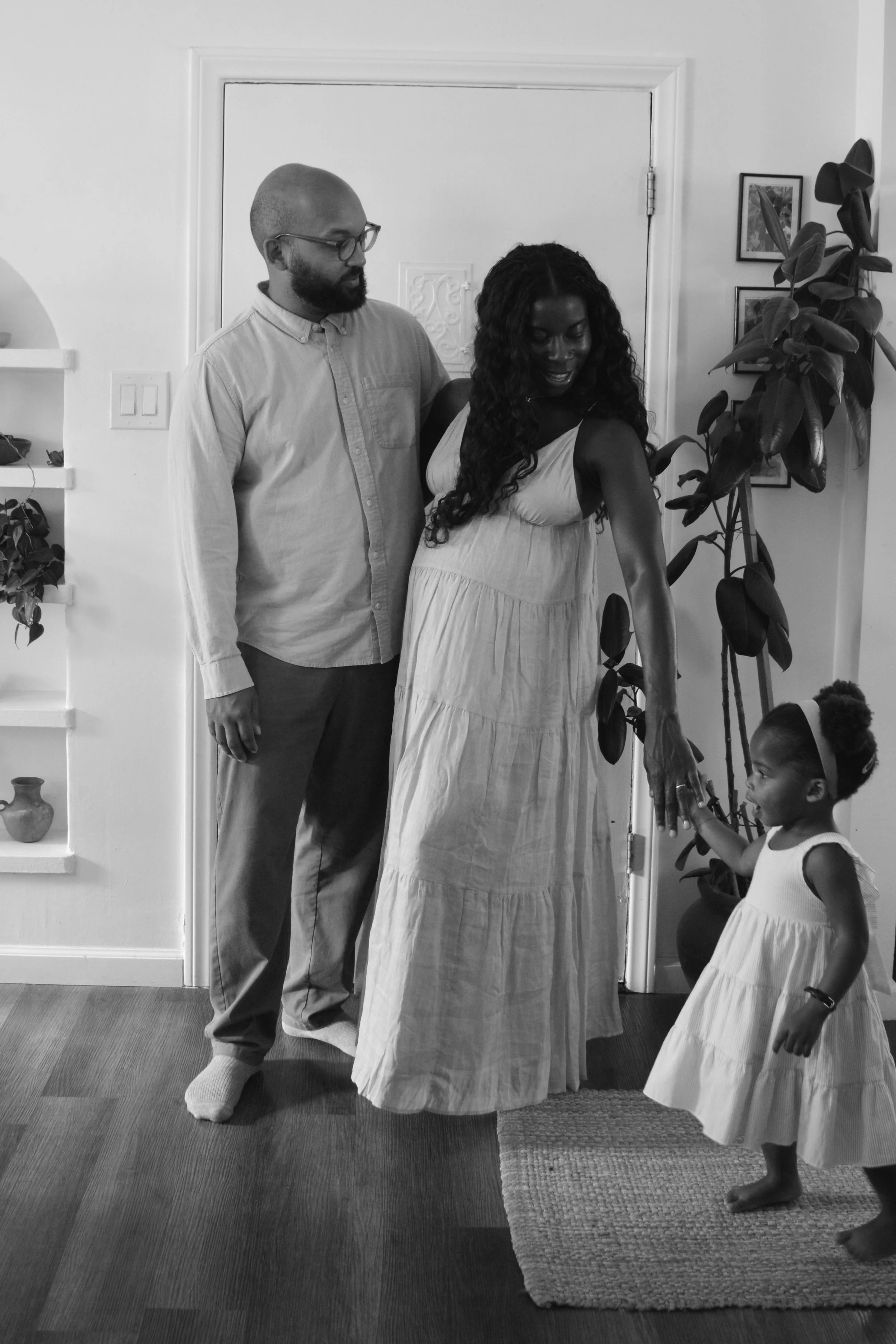 A black-and-white photo of a man with glasses and a beard, standing beside a woman with long curly hair, who is wearing a light dress. They are in a room with a white wall, artwork on the wall, a large potted plant, and a child in a light dress holdi