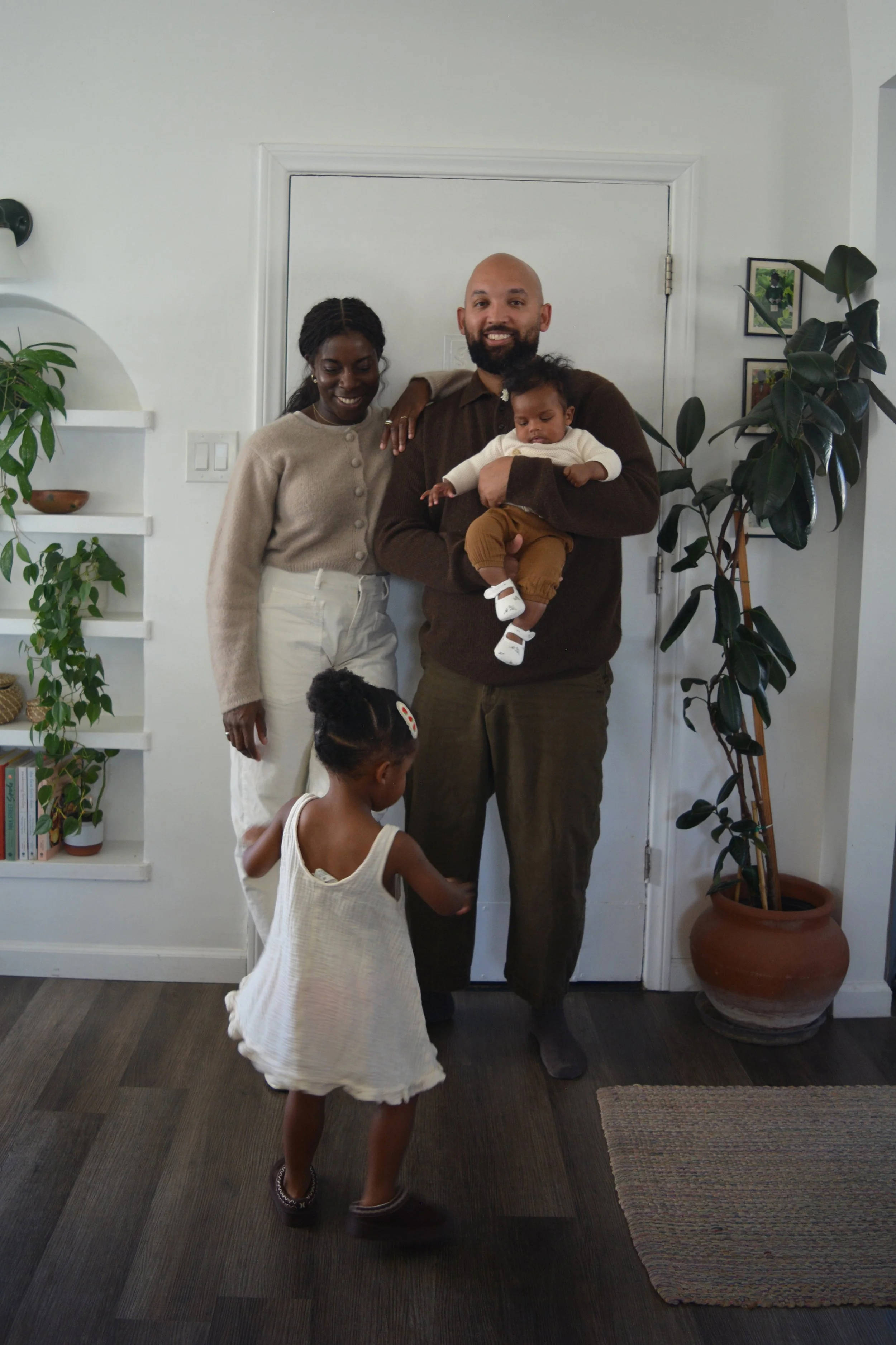 A family moment with two adults and two children inside a modern home, with one adult holding a baby and the other with a hand on the baby's shoulder, smiling at the camera, surrounded by indoor plants and framed pictures on the wall.