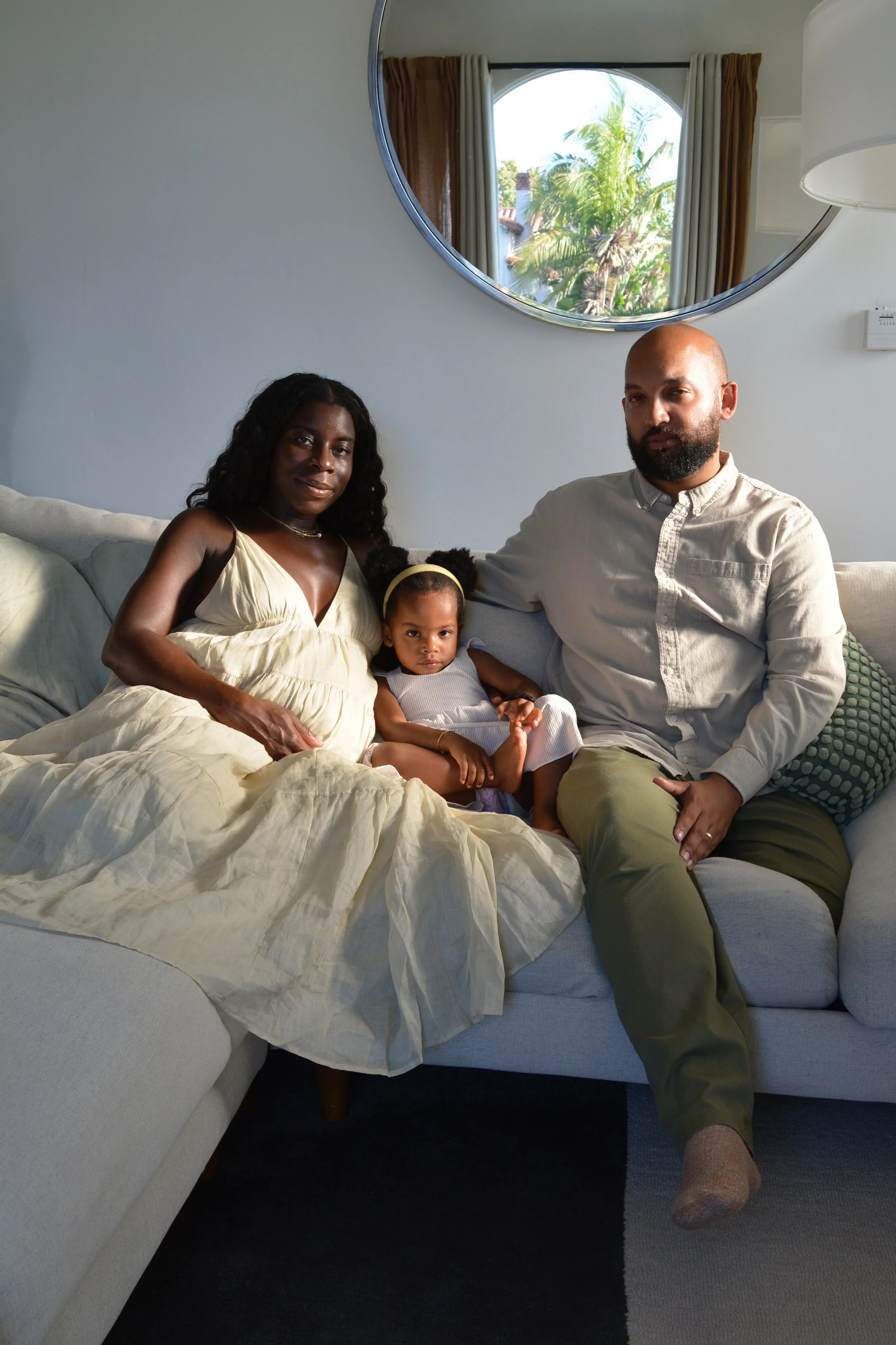 A family of four sitting on a beige couch in a living room, with a large round mirror on the wall above. The room has a window with a view of palm trees and blue sky outside. The family appears relaxed, with the adults smiling and the child sitting b