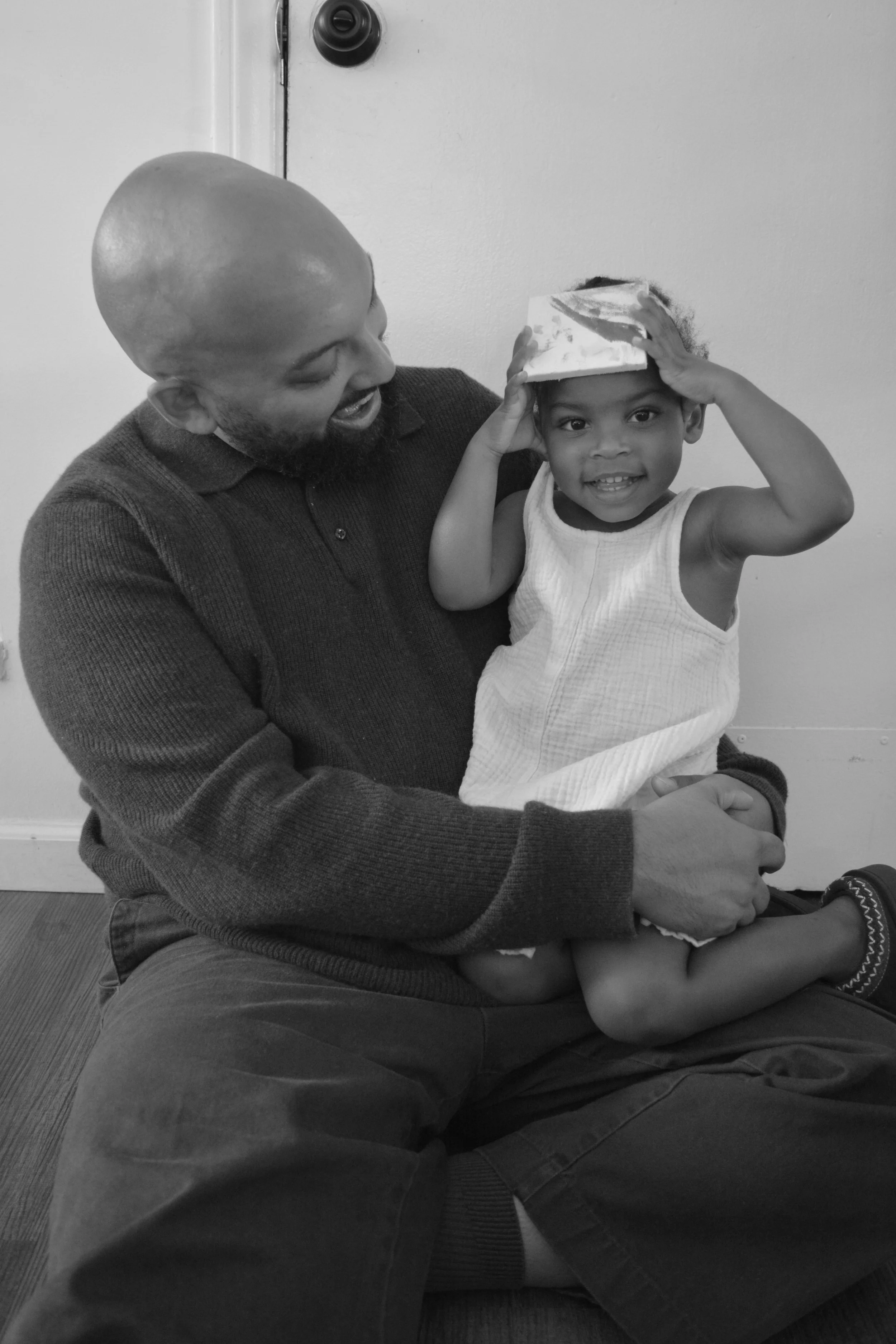 A man and a young girl, sitting on the floor against a plain wall, sharing a joyful moment. The girl is smiling and playfully tilting a square object, possibly a book or a box, on her head while the man looks at her with affection.