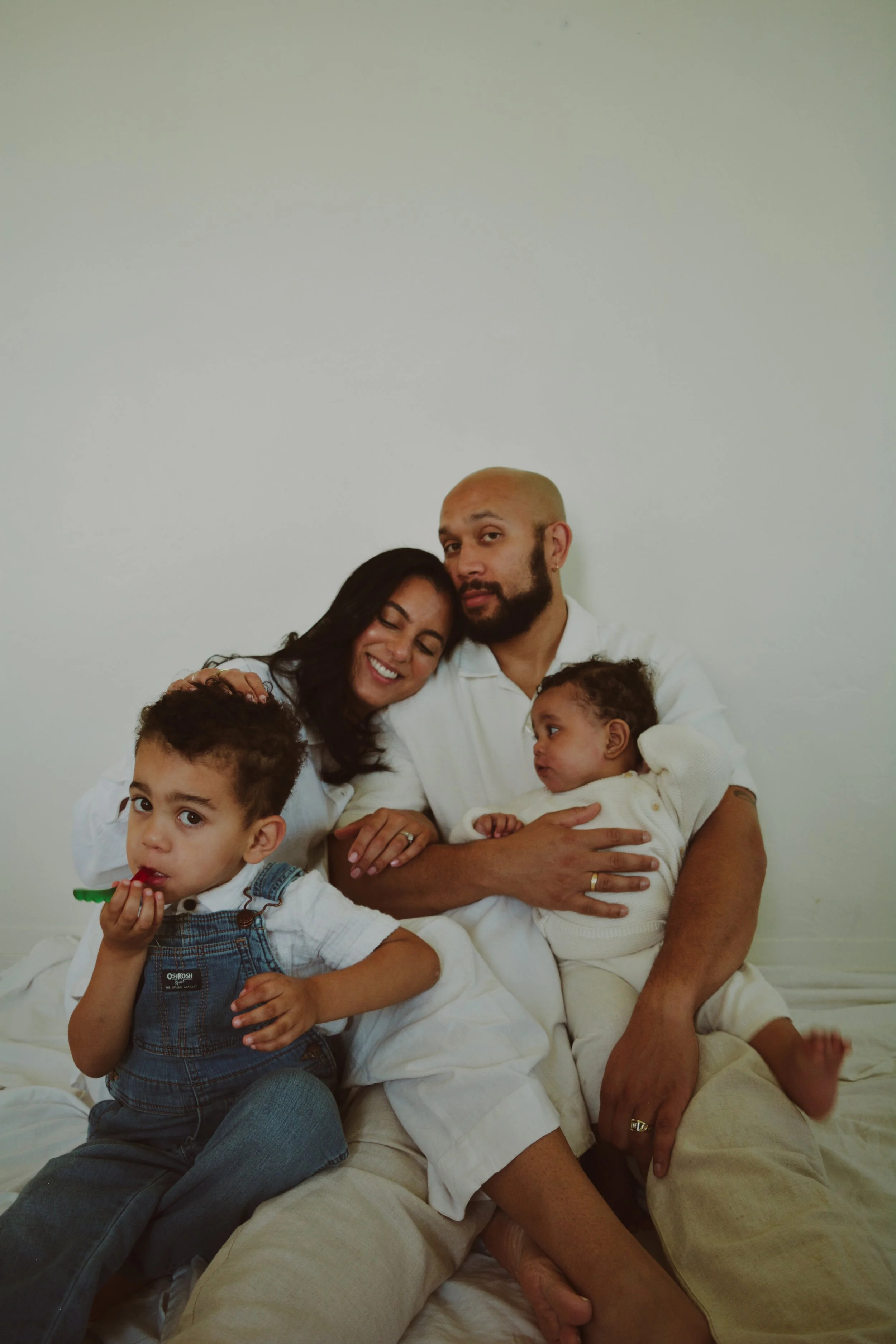 A family of five sitting on a bed against a plain white wall, including a man, a woman, and three children. The woman is smiling with her eyes closed, resting her head on the man's shoulder. The man is holding a baby and looking at the camera. One ch