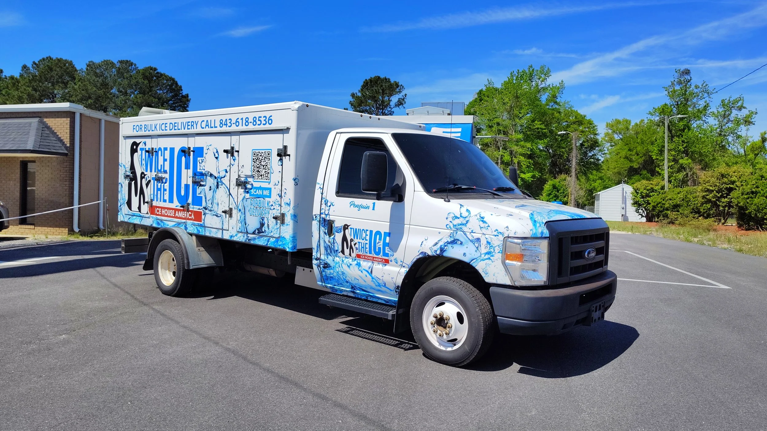 White ice delivery truck with blue and black branding parked on a paved lot. Trees and a small building are in the background, under a clear blue sky.
