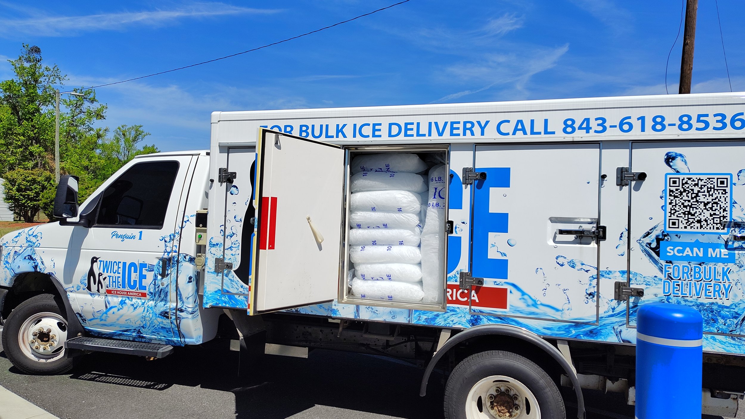White delivery truck with blue ice graphics, open door revealing bags of ice inside, parked on a street with trees and utility pole in background.