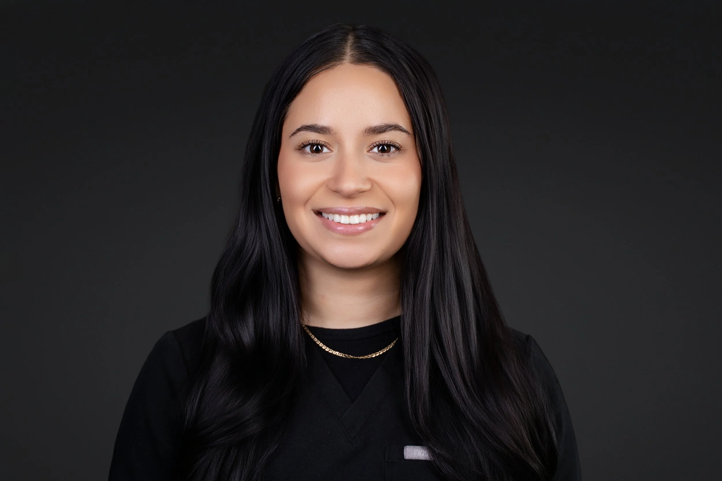 Portrait of a smiling woman with long dark hair, wearing a black top and gold necklace, against a dark background.