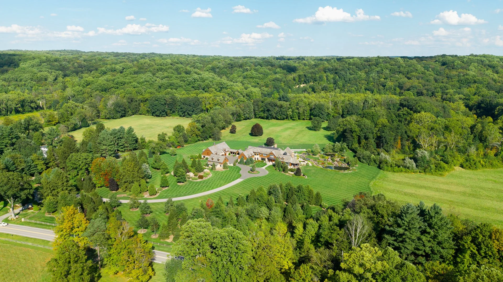 A large estate surrounded by lush green trees and landscaped gardens in a rural area with open fields and a wooded hillside in the background, under a partly cloudy sky.