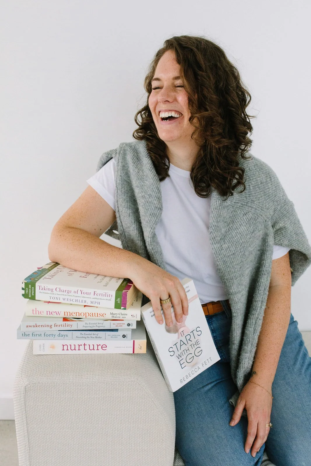 A woman laughing and leaning on a stack of books. She has curly brown hair, light skin, and is wearing a white t-shirt with a gray sweater draped over her shoulders. She is holding a book titled 'It Starts with the Egg' by Rebecca Fett.