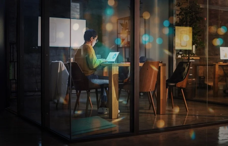 Persona sentado en una cafetería o espacio de coworking, trabajando en una laptop, vista a través de una ventana de vidrio en la noche, con luces de la ciudad difusas en el fondo.