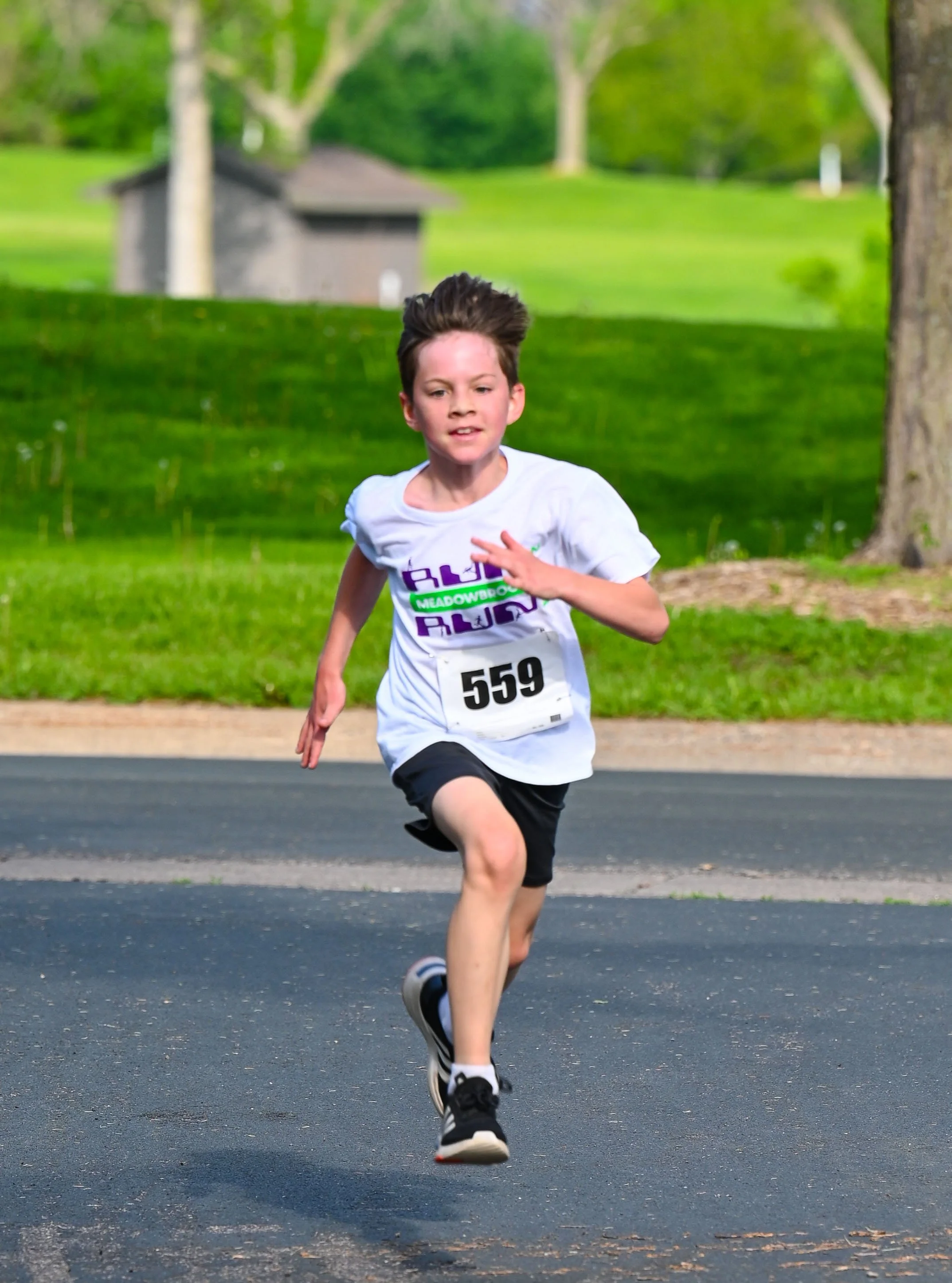 A young boy running in a race outdoors in a park, wearing a white shirt with purple and green logo, black shorts, and a race number 559.