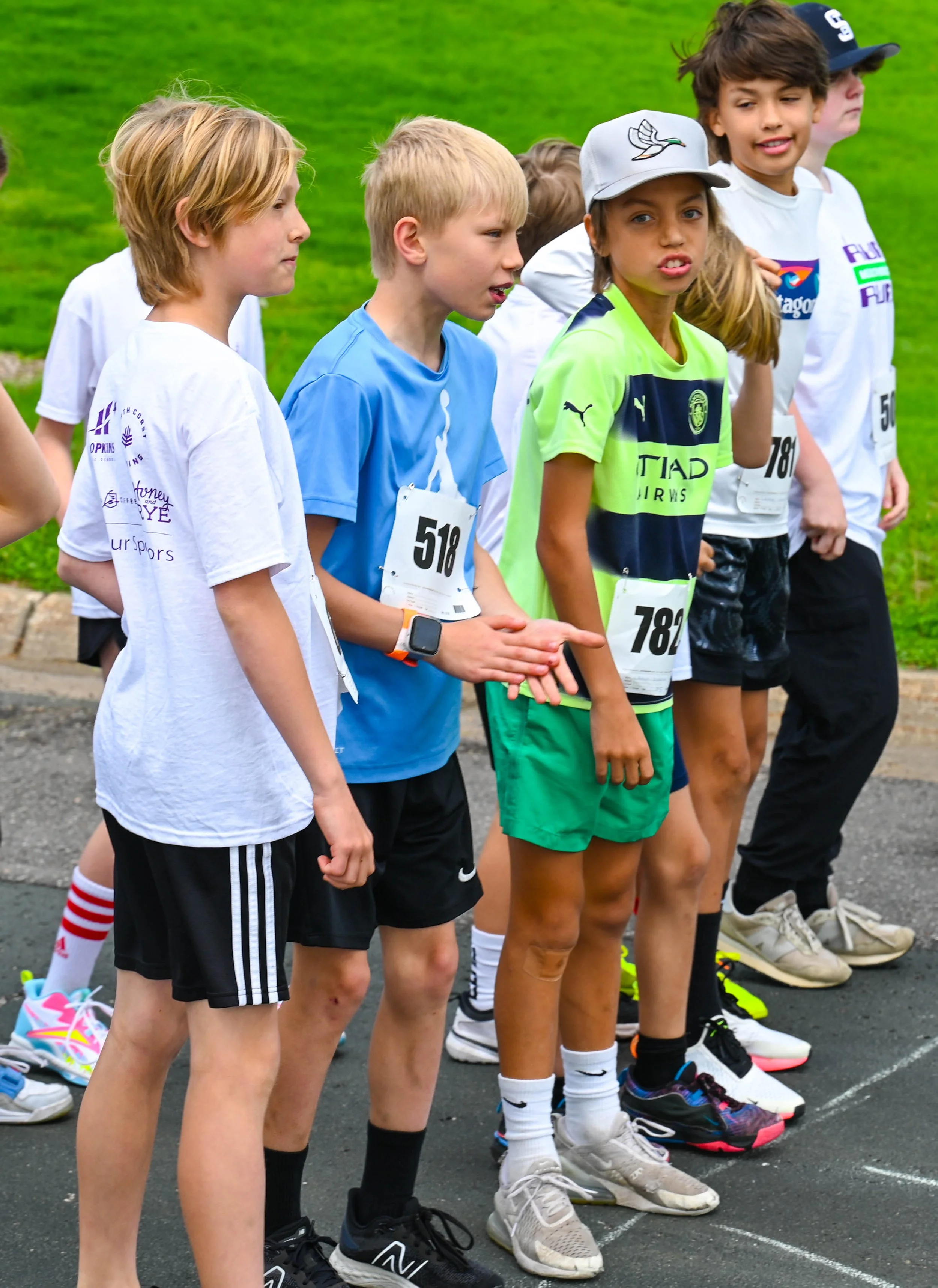 Children in athletic clothing and race bibs lined up at a starting line for a race, standing outdoors on a paved surface with a grassy field in the background.