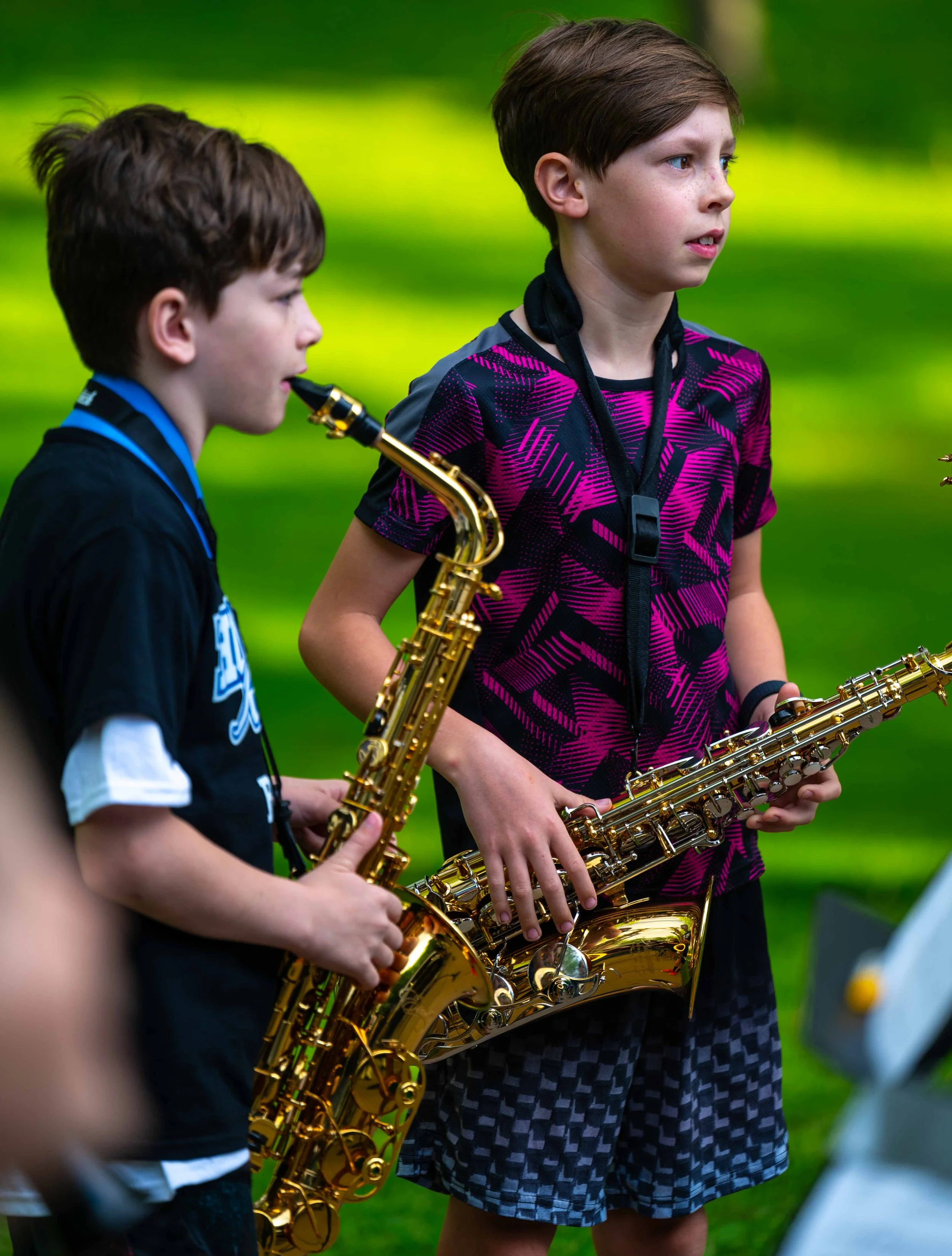 Two boys holding saxophones in an outdoor setting, greenery in the background.