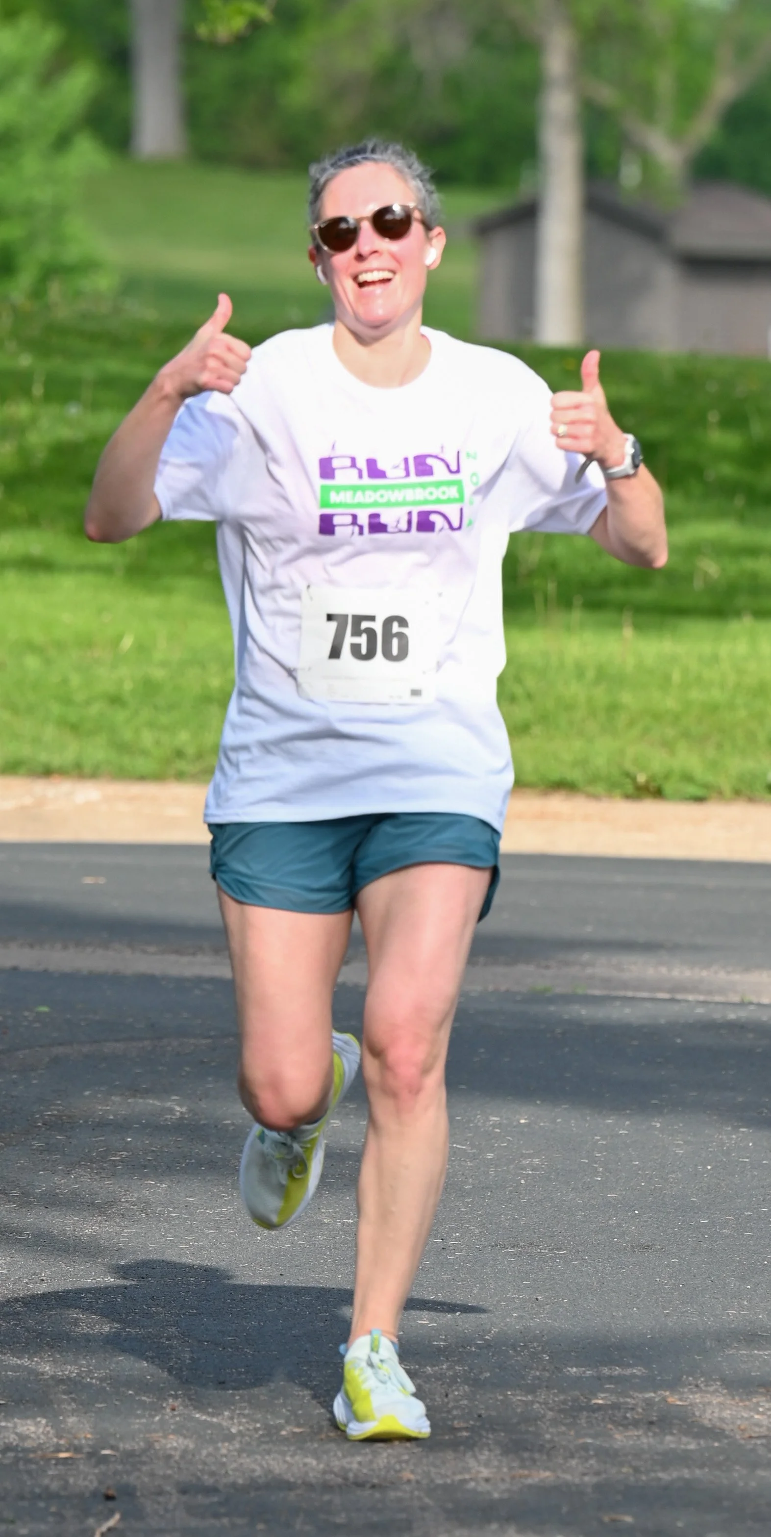 A woman running in a race, smiling and giving a thumbs-up, wearing sunglasses, a white race bib with number 756, a white T-shirt, teal shorts, and yellow and white running shoes, on a paved path with green trees in the background.