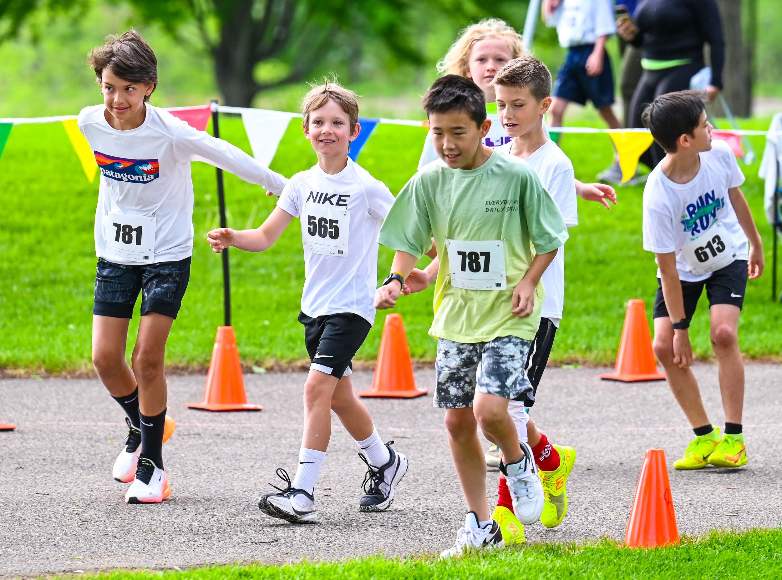 Children participating in a running race on a pathway marked with orange cones in a grassy park. The kids are wearing race bibs with numbers.