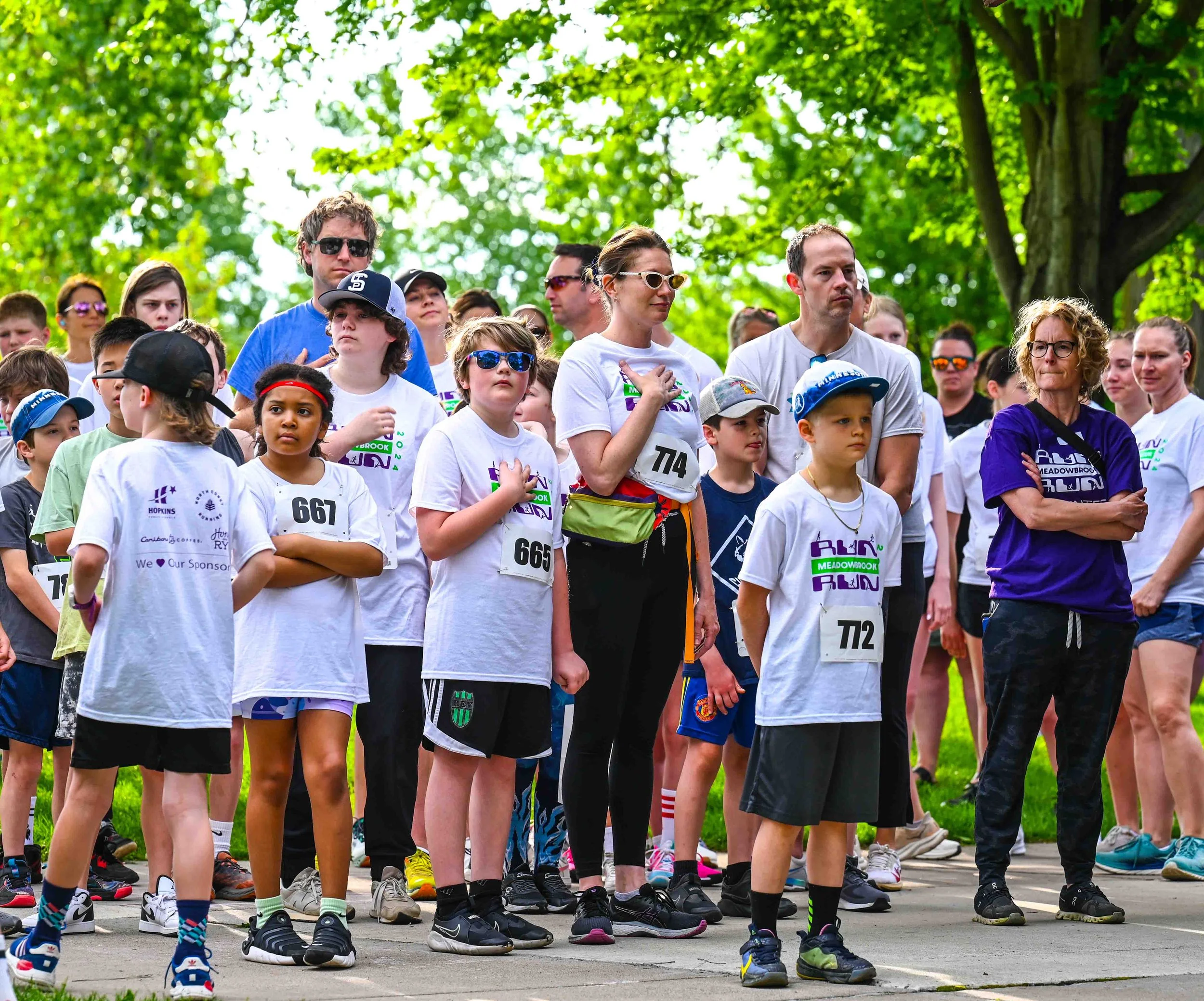 Group of children and adults gathered in a park, some with race bibs, ready for a run event on a sunny day with green trees in the background.
