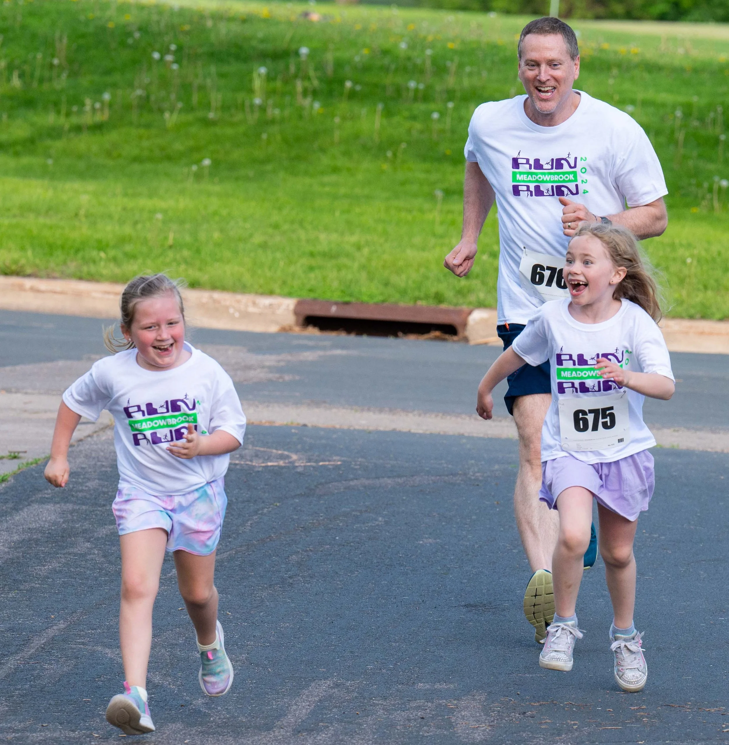 Two young girls and an adult male participating in a fun run, running on a paved path with a grassy field in the background. All are wearing white t-shirts with race logos and numbered bibs, smiling and enjoying the event.
