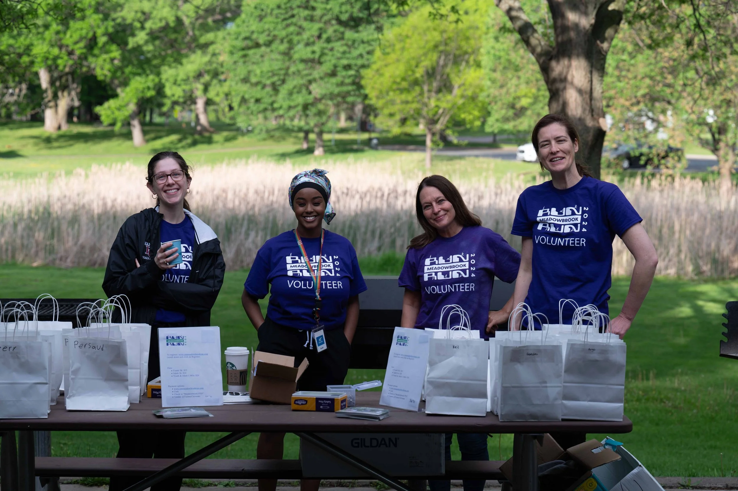 Four women wearing matching blue volunteer t-shirts standing behind a table with bags and supplies outdoors in a park with green trees and grass.