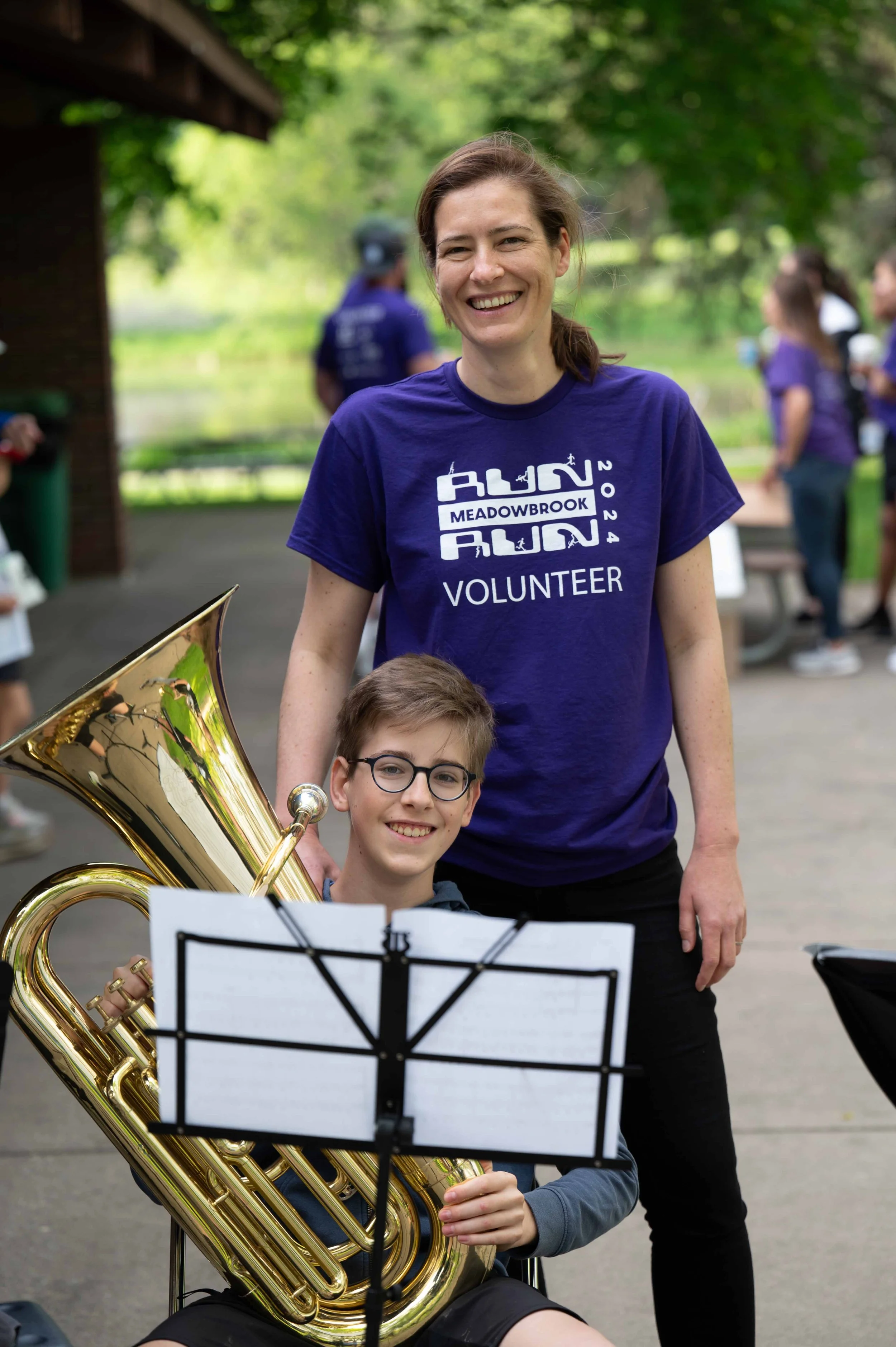 A smiling woman in a purple volunteer t-shirt standing next to a boy with glasses playing a large brass instrument, possibly a euphonium, outdoors with people in the background.