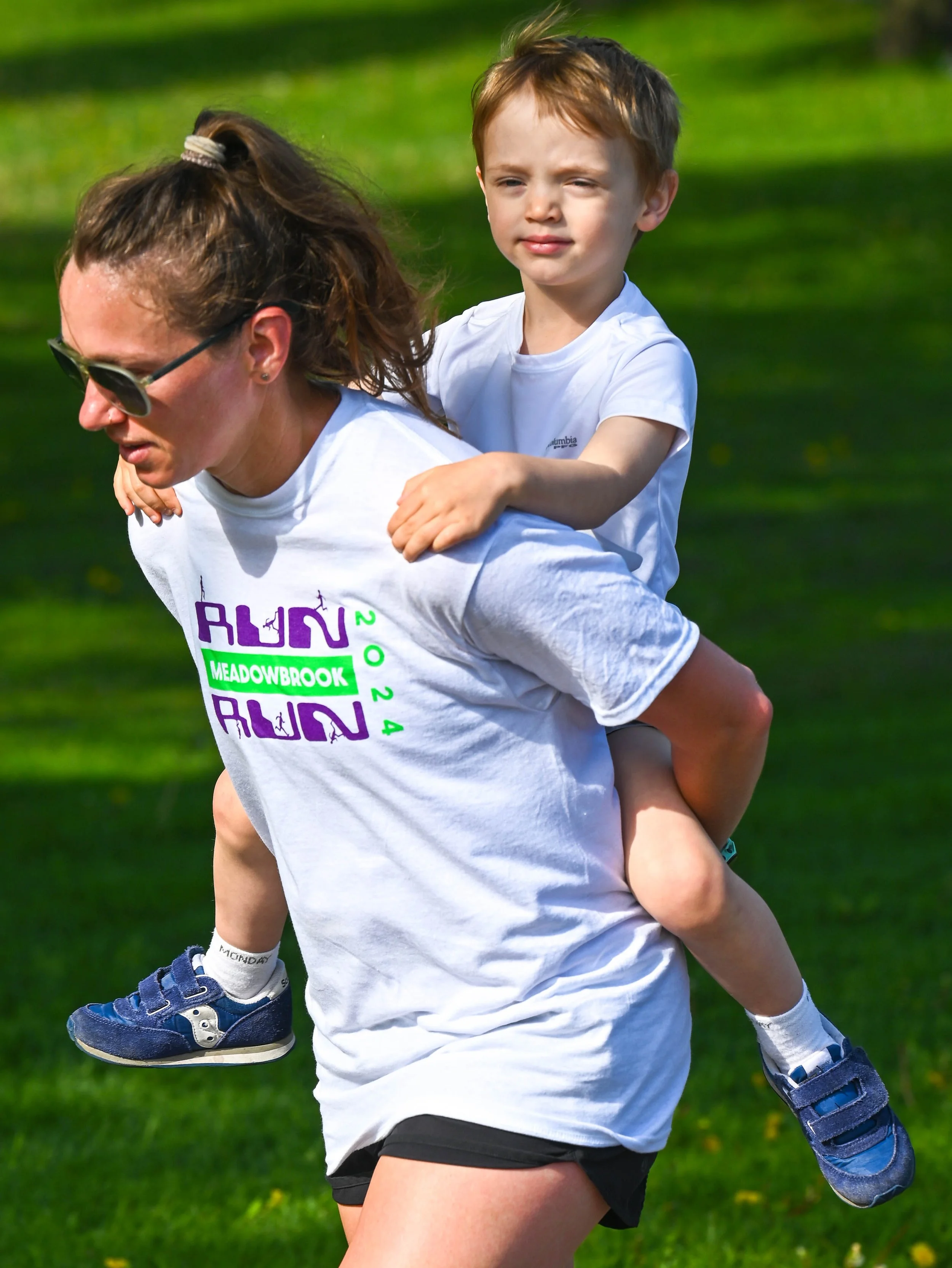 A woman is running outdoors with a young boy on her back, both wearing white T-shirts. The woman has sunglasses and a ponytail. The boy is holding onto her shoulders and is looking ahead.
