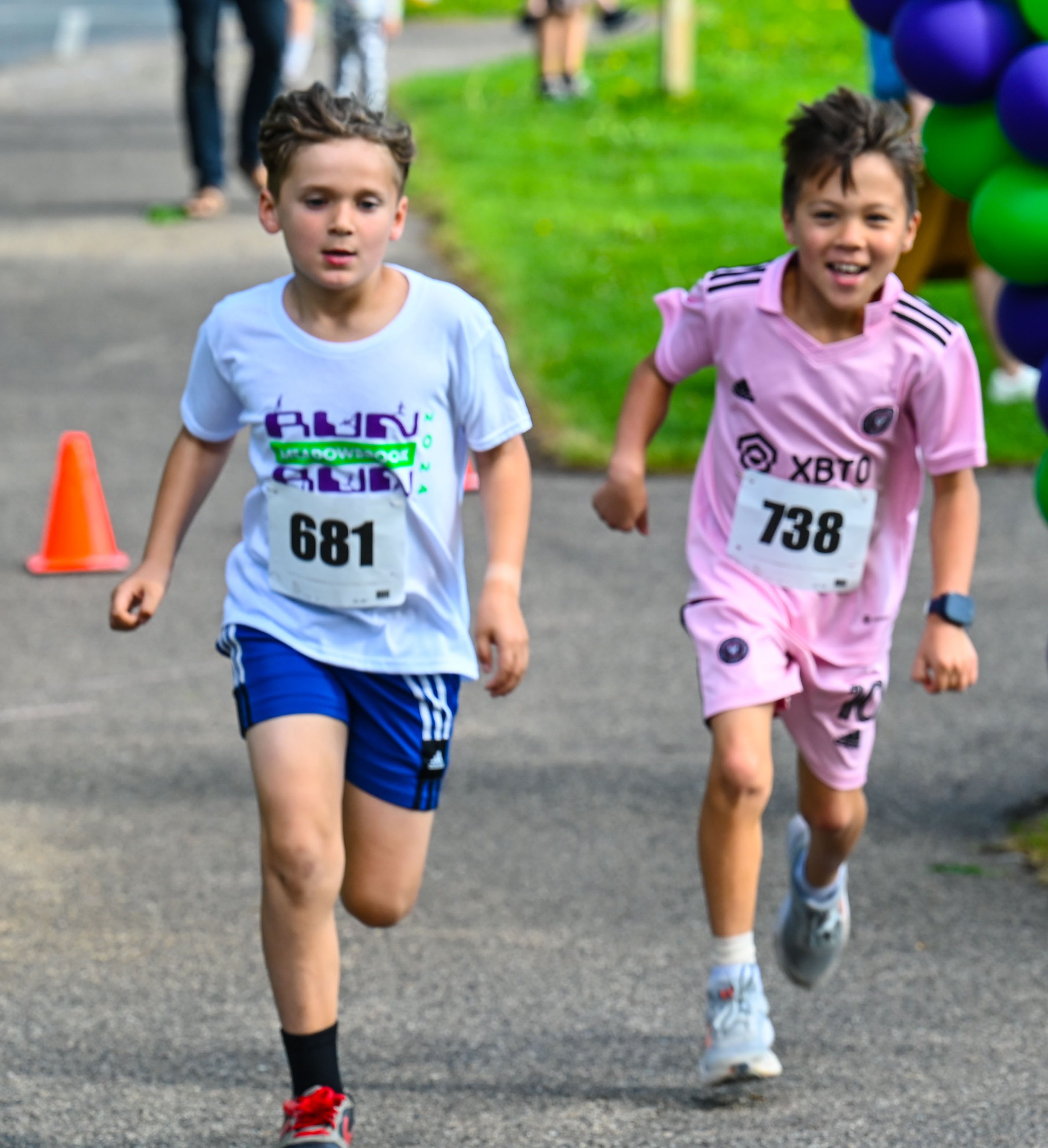 Two boys running in a race, wearing numbered bibs, with the boy on the left in a white shirt and blue shorts, and the boy on the right in a pink outfit, on an outdoor running track with orange cones and balloons in the background.