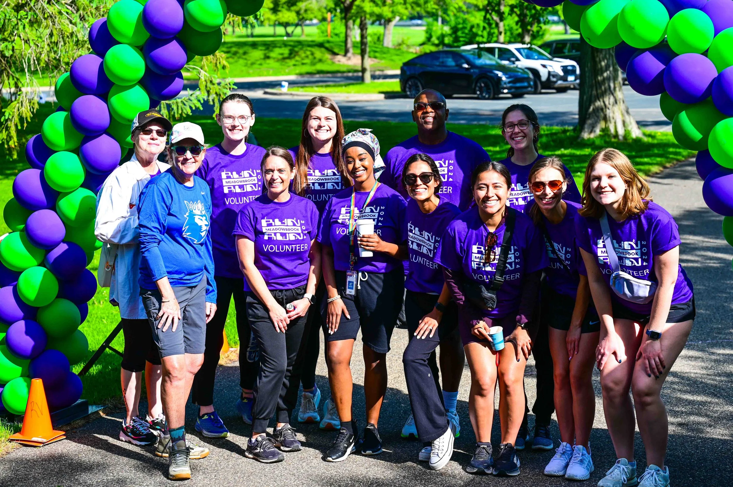 Group of volunteers posing under a purple and green balloon arch at an outdoor event, some wearing purple shirts with event branding.
