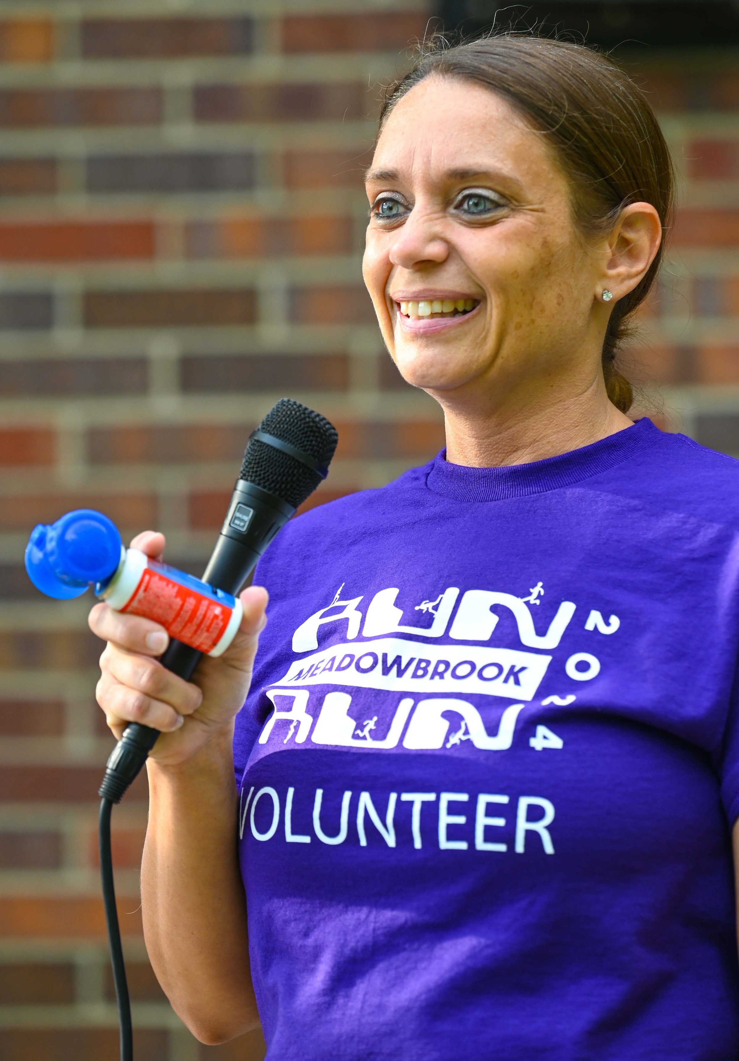 A woman speaking into a microphone during an outdoor event, wearing a purple volunteer T-shirt that says 'ALV Meadowbrook 2014 Volunteer,' holding a container of bubbles, with a brick wall in the background.