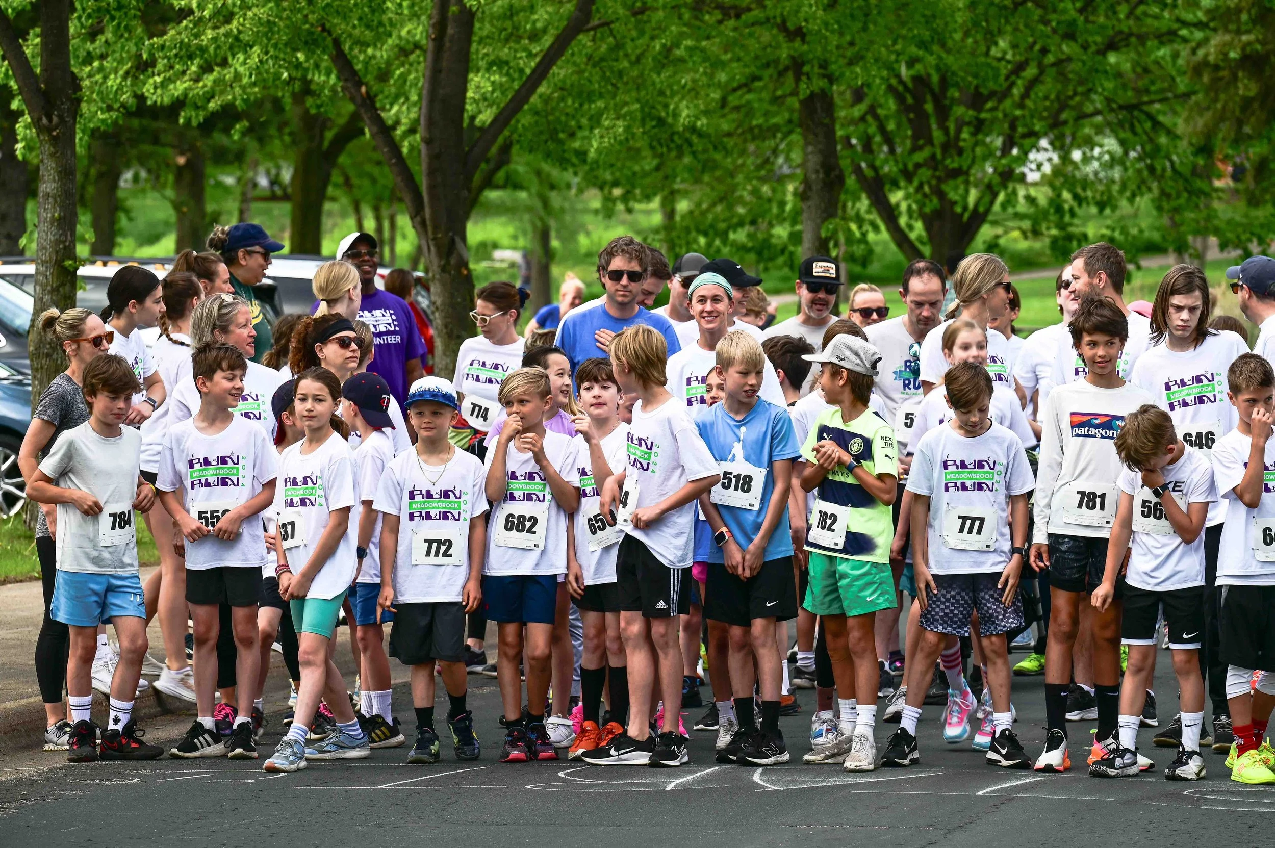 Group of children and adults at the starting line of a race in a park with green trees in the background.