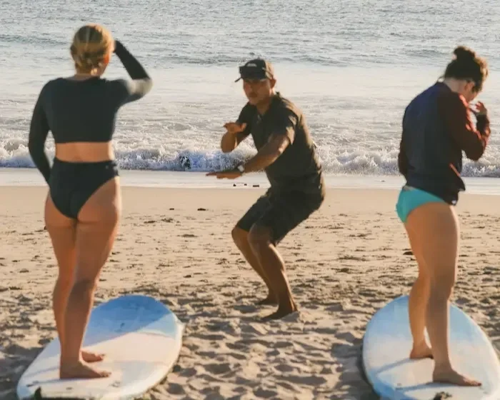 Two women with surfboards and a man on a beach near the water, with the man filming or taking a picture of the women.