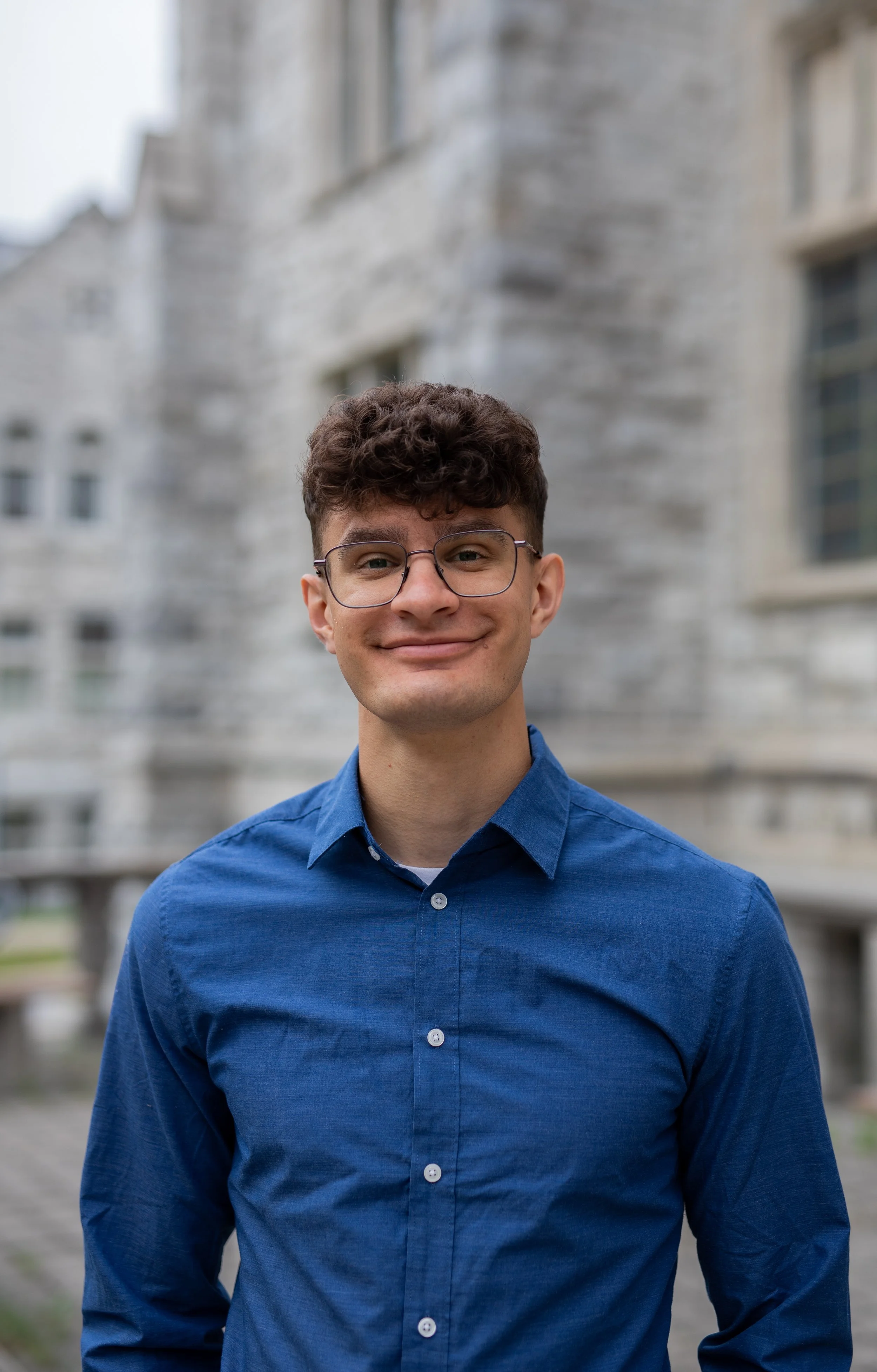Young man with brown curly hair and glasses smiling, wearing a blue shirt, standing outdoors with a blurred stone building in the background.