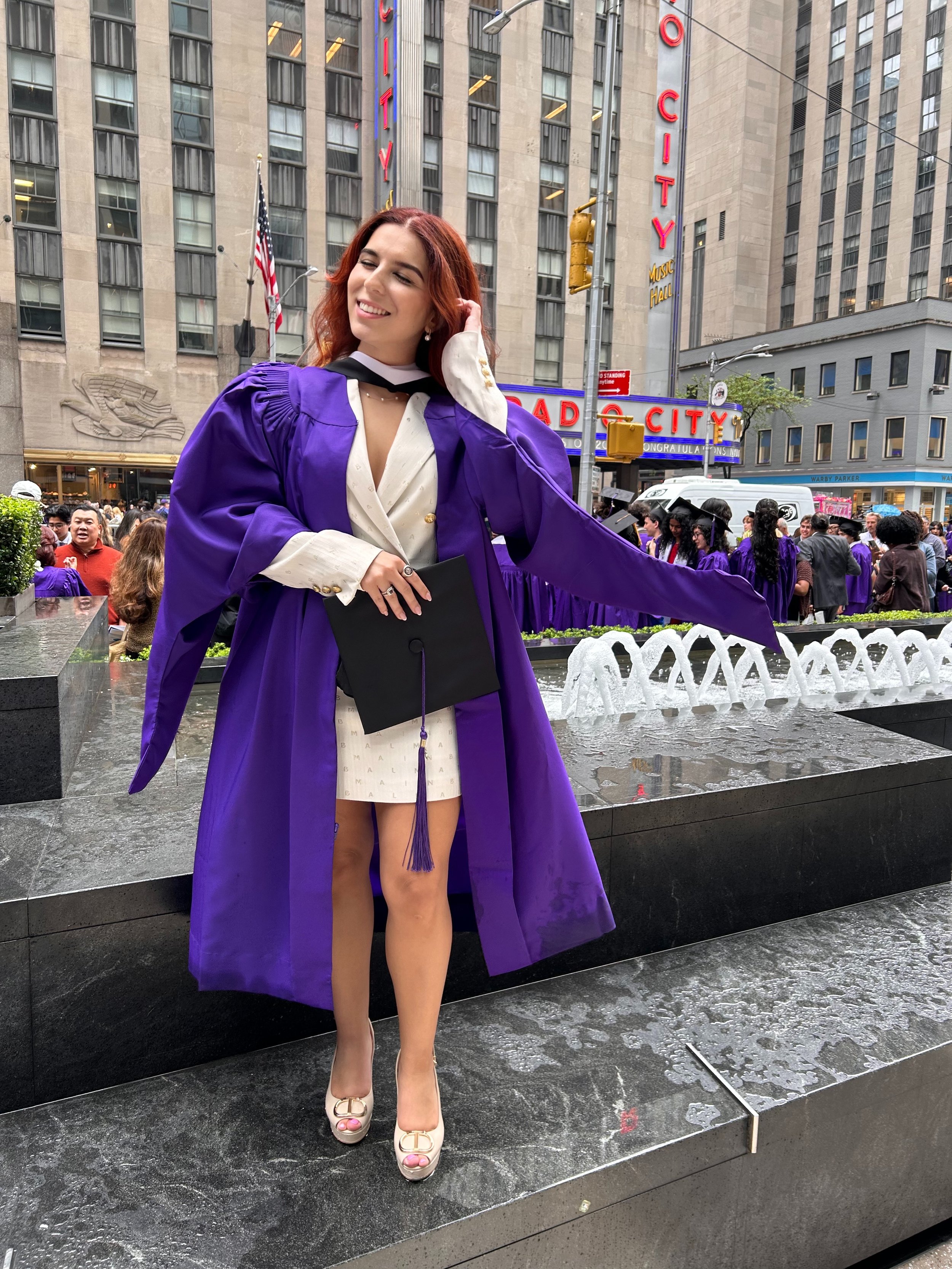 Young woman in a purple graduation gown and cap, holding a black diploma folder, standing outside at a graduation ceremony in front of Radio City Music Hall in New York City, with a fountain and other graduates in the background.