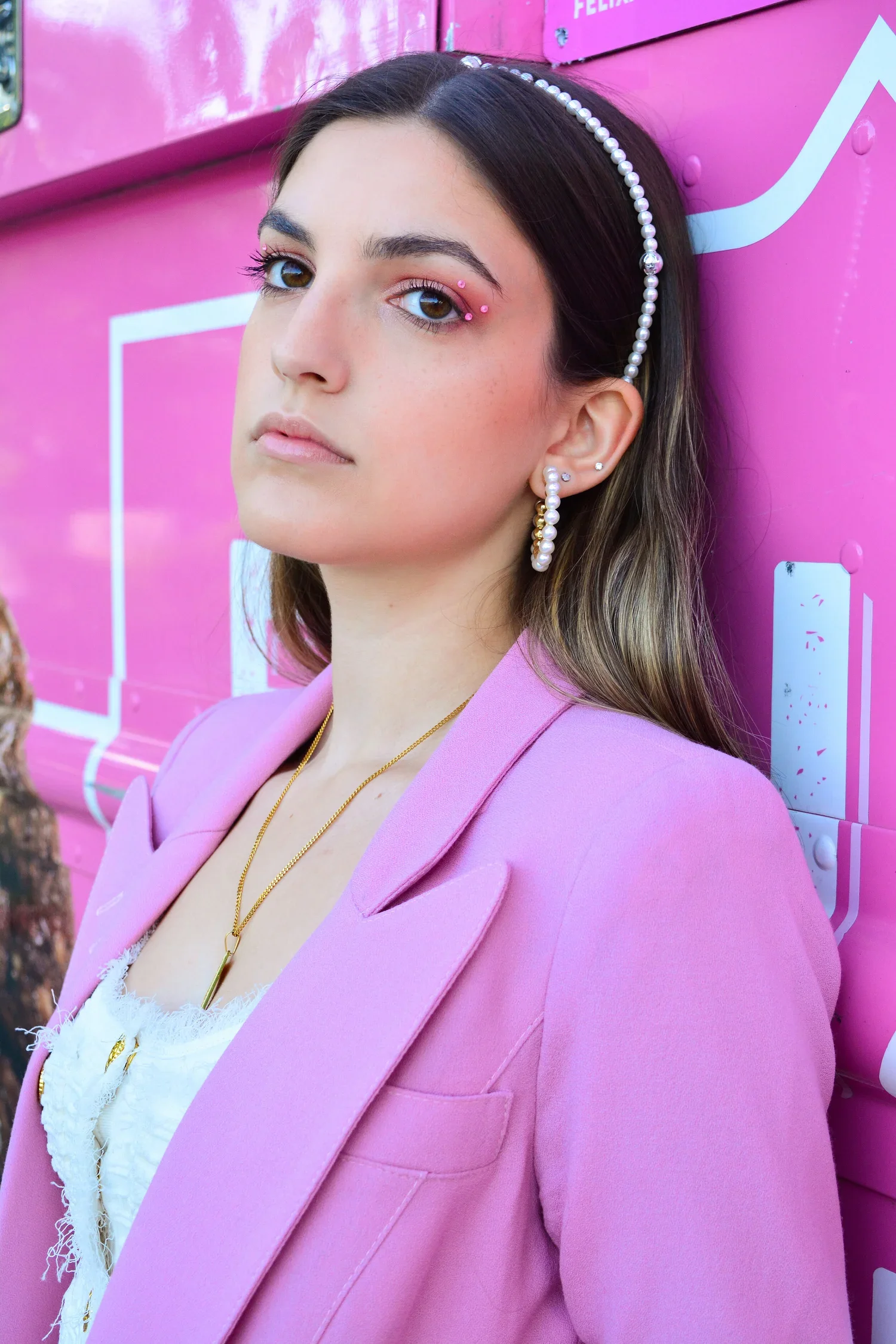 A young woman with dark hair, wearing a pink blazer, gold necklace, and pearl earrings, stands against a pink background with white and blue accents.