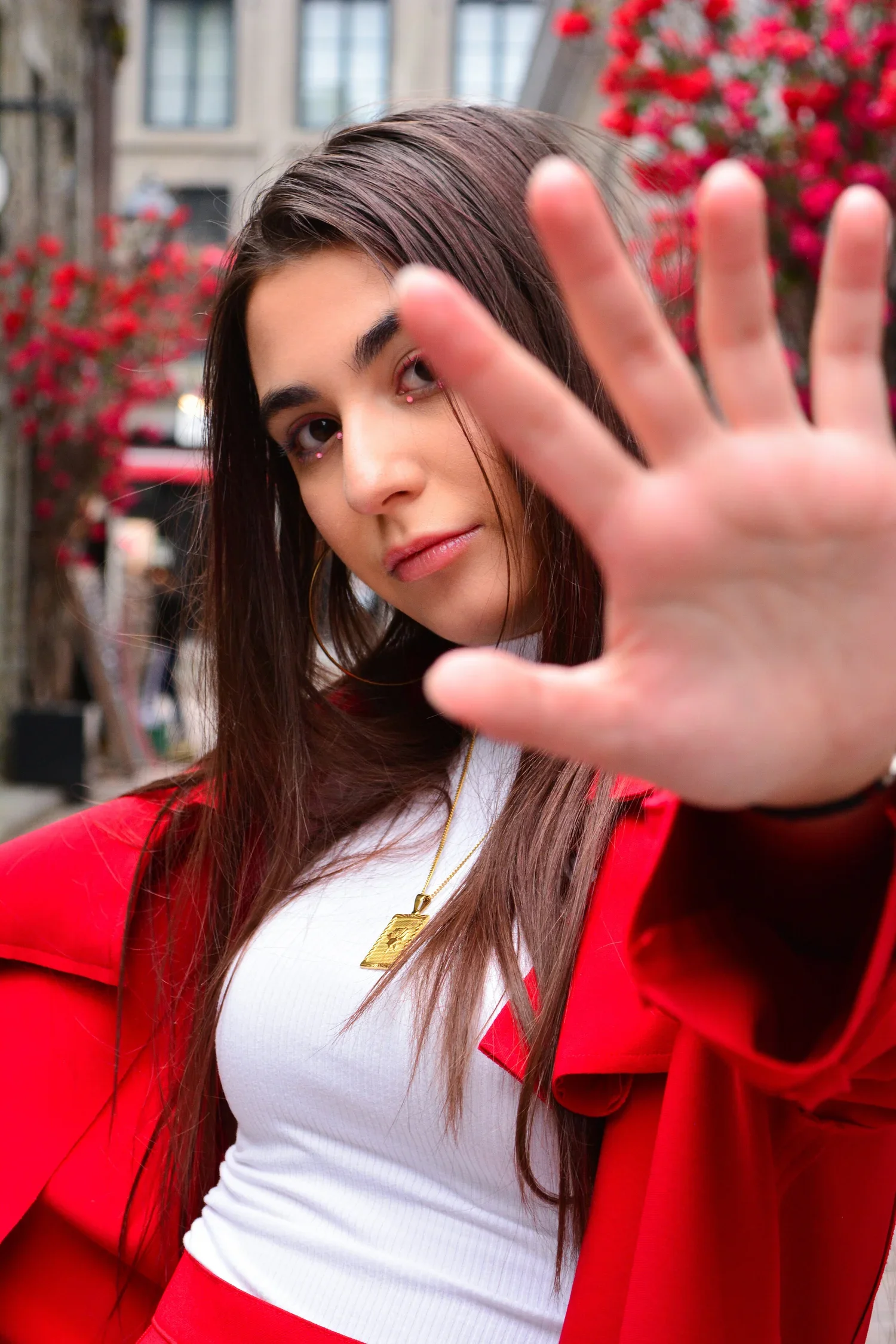A young woman with long brown hair wearing a red coat and white top, raising her hand toward the camera in an outdoor setting with buildings and pink flowering trees in the background.