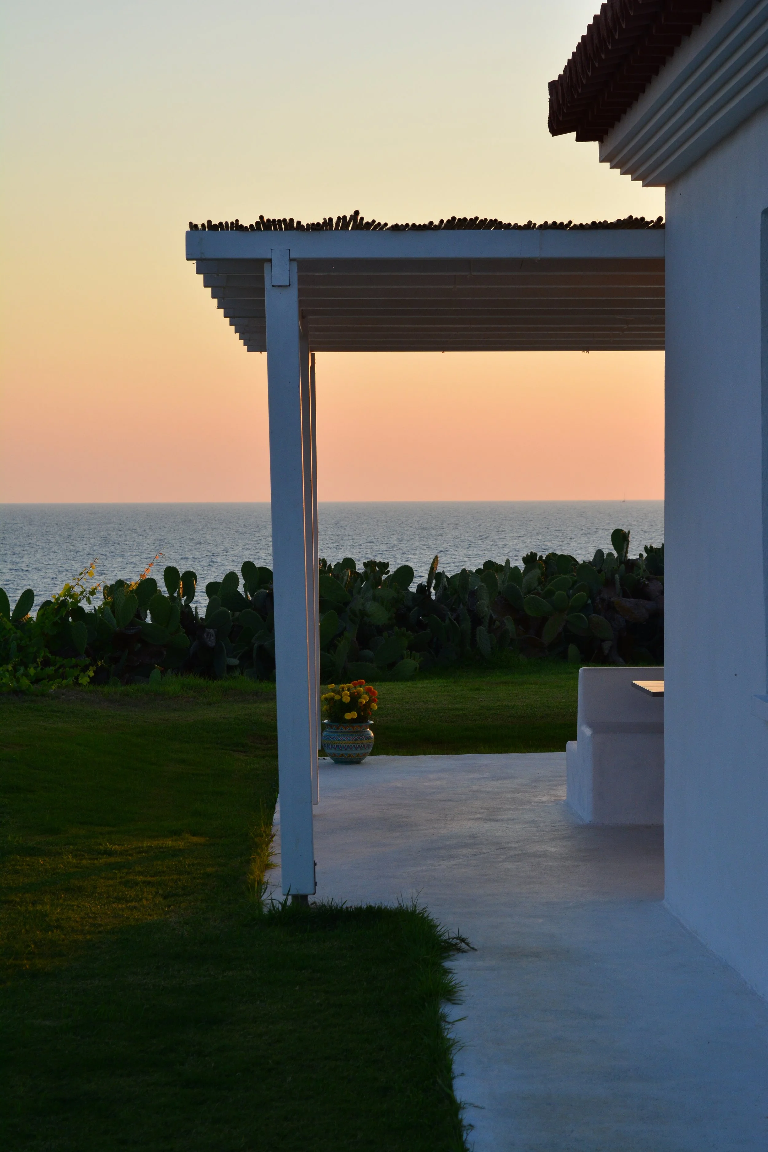 Beach house patio with white walls, green grass, potted plant, and ocean view during sunset.