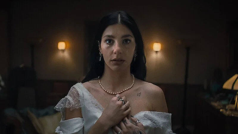 A woman in a white dress with lace details, wearing pearl jewelry, standing in a dimly lit room with wall lamps, looking directly at the camera.