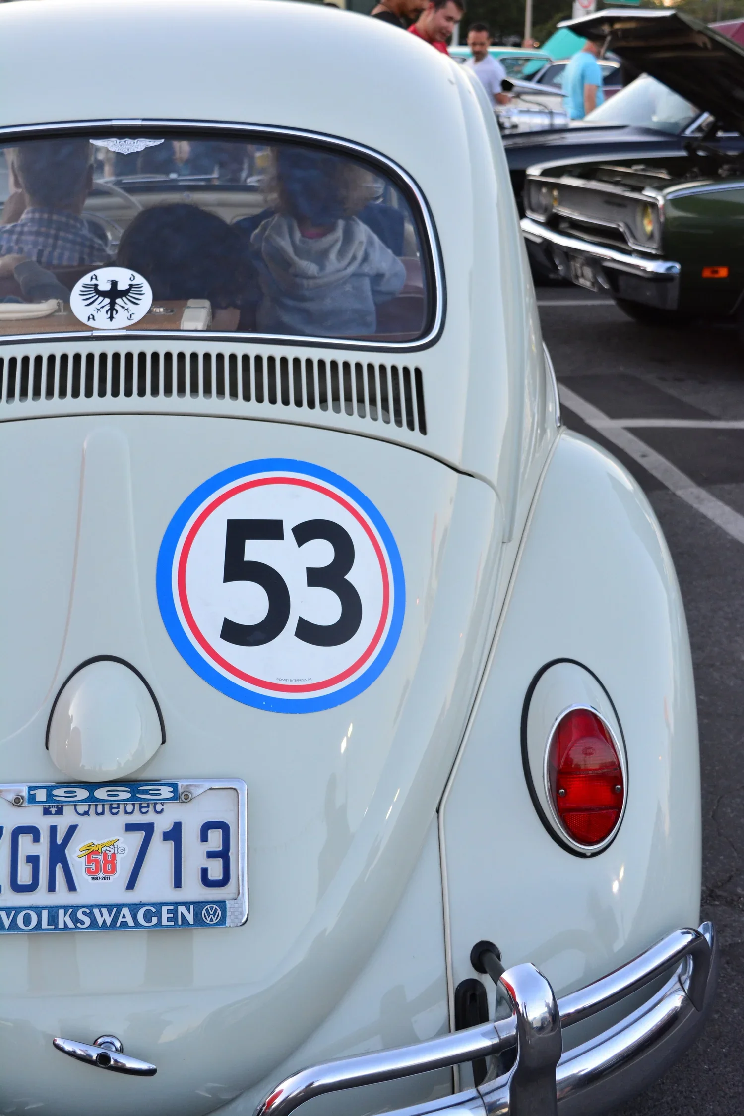 Rear view of a white vintage Volkswagen Beetle with a number 53 decal on the back, parked at a car show, with people and other classic cars in the background.