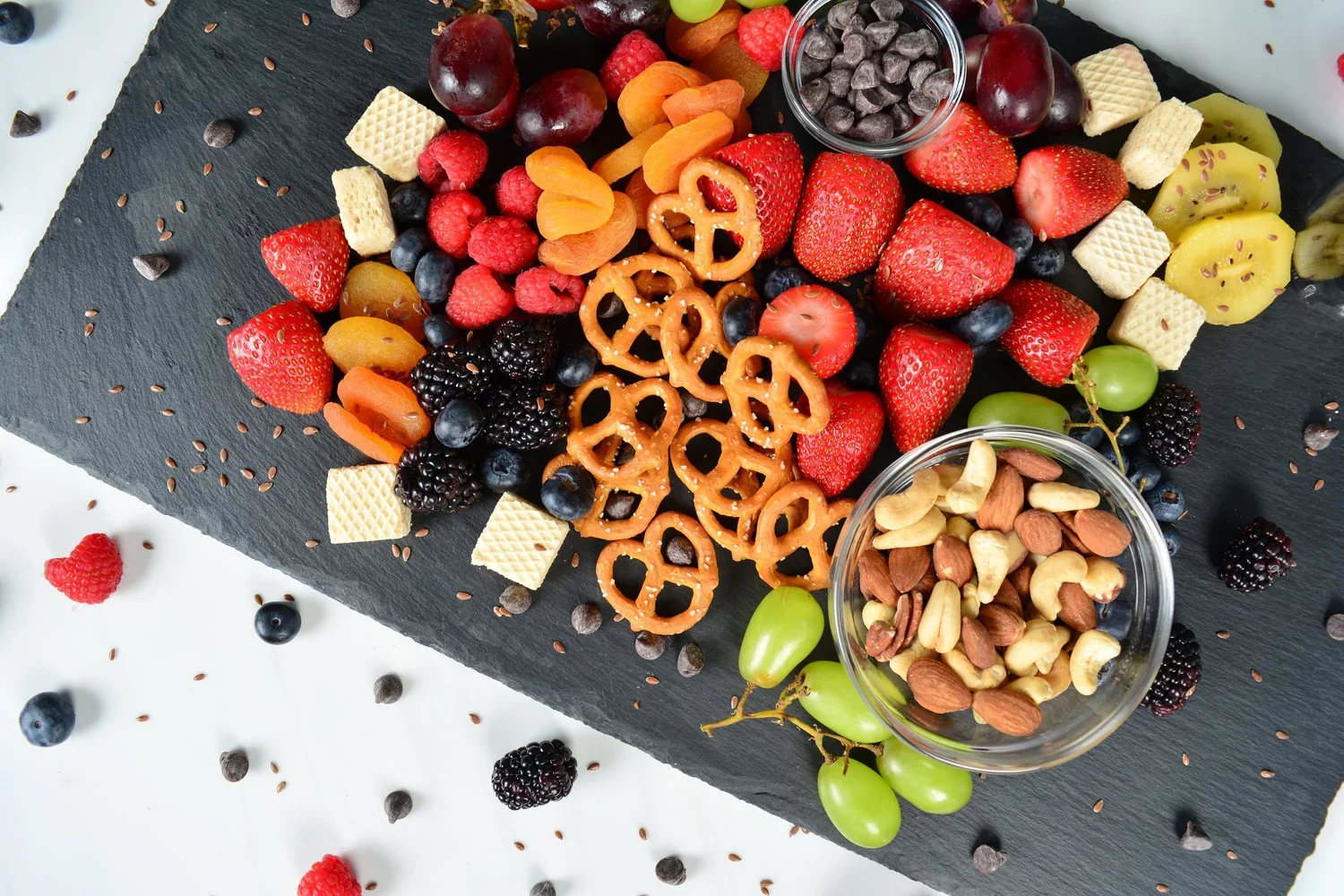 A black slate platter filled with an assortment of fresh berries, sliced kiwi and strawberries, mixed nuts, pretzels, mini wafers, and small bowls of chocolate chips and dried apricots.