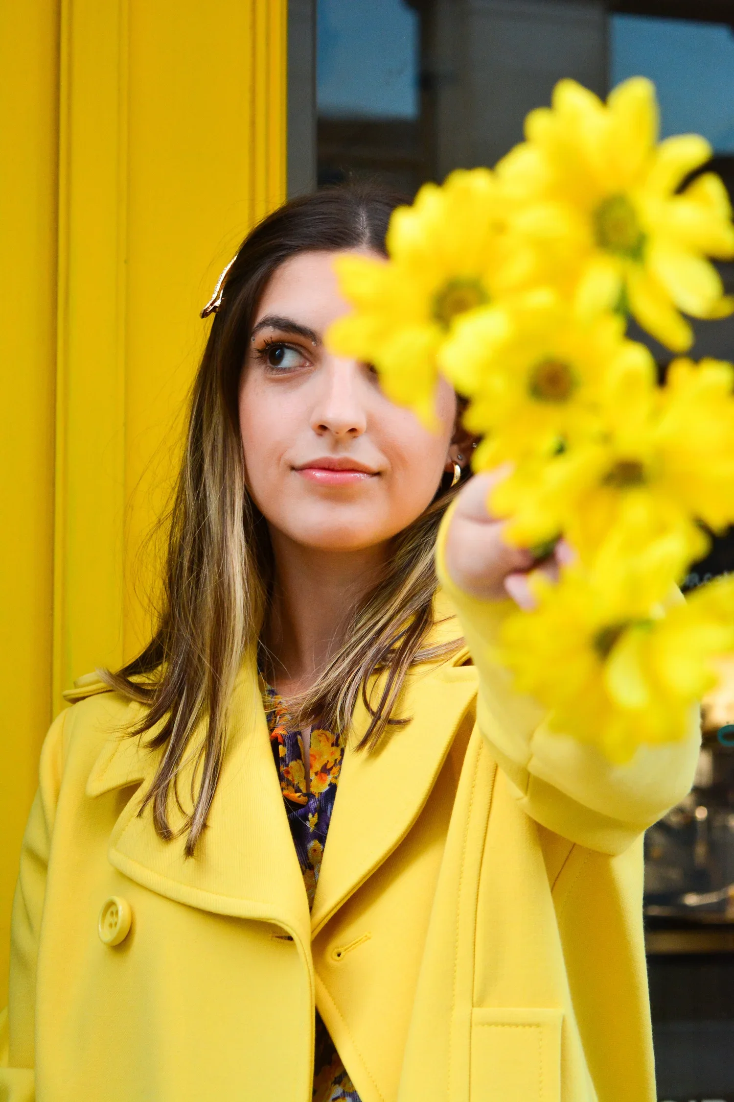 Woman in a yellow coat holding a bunch of yellow flowers, standing against a yellow wall.