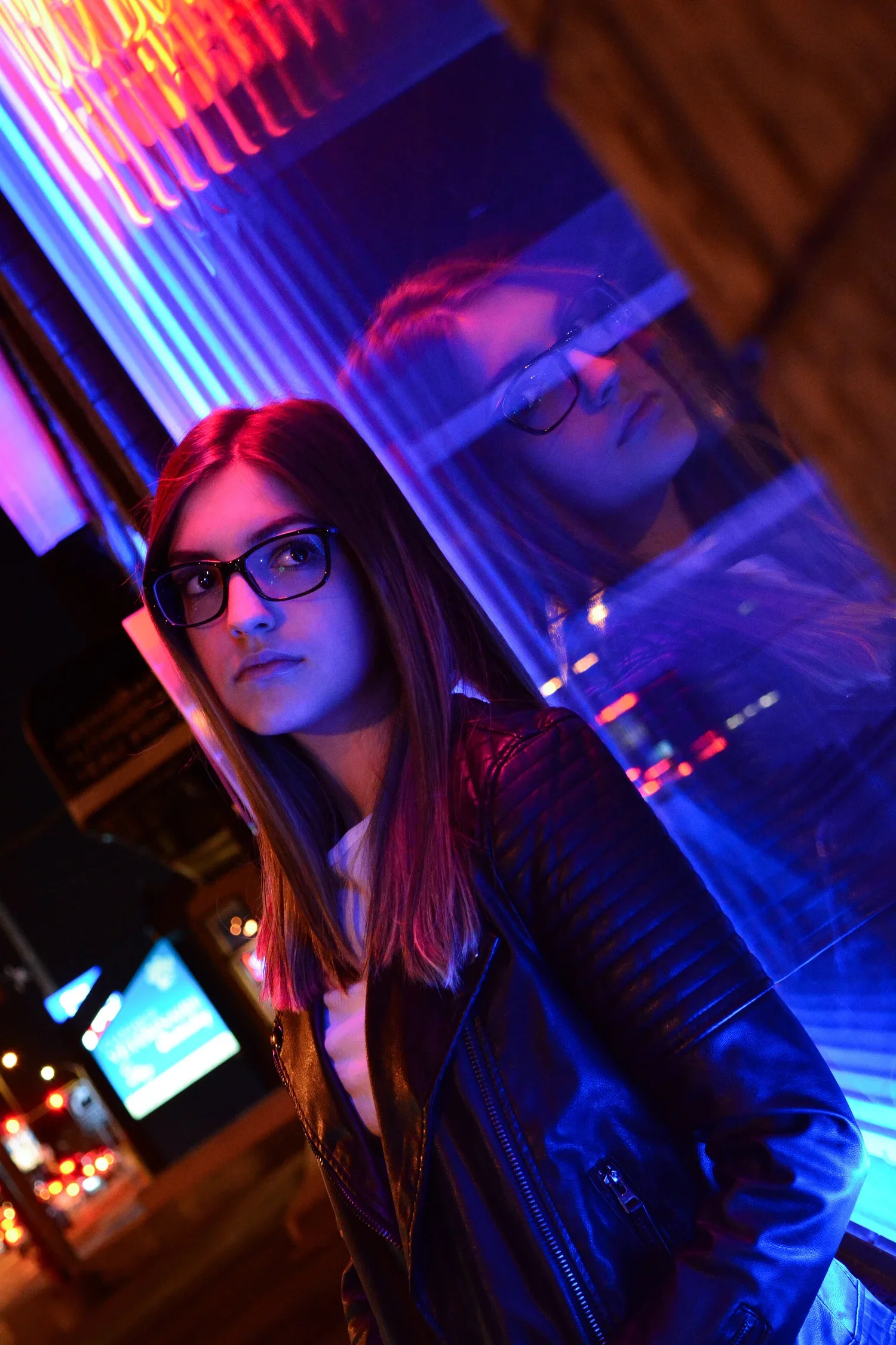 A young woman with glasses and long hair stands at night next to a reflective glass surface, illuminated by vibrant neon lights with city street in the background.