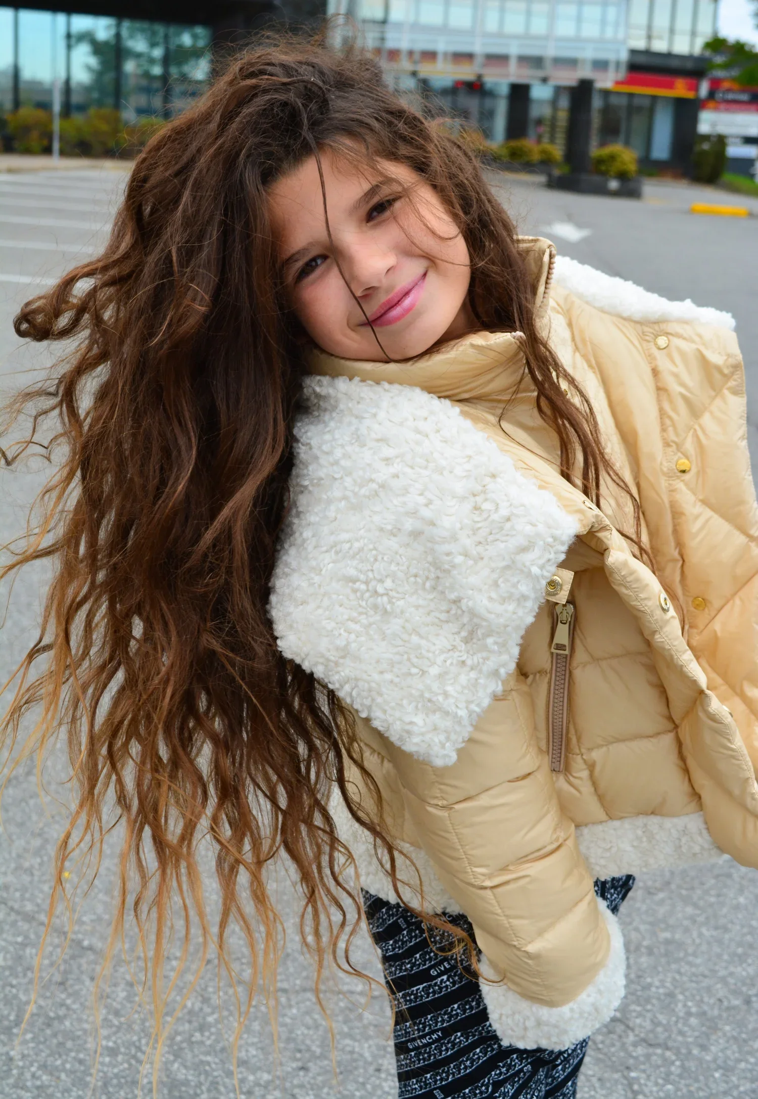 A young girl with long, curly brown hair smiling and leaning sideways in an outdoor parking lot with a glass building in the background. She is wearing a beige jacket with a white, fluffy lining.