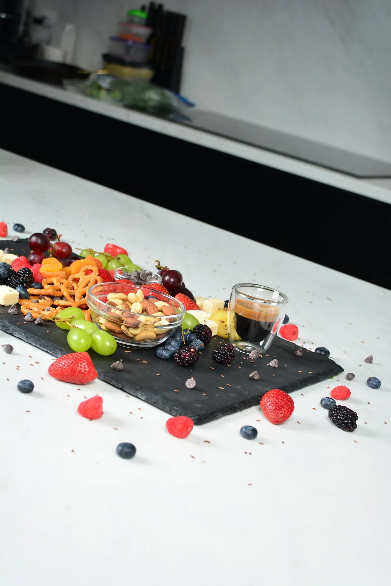 Assorted fresh fruit, nuts, cookies, and espresso on a black slate serving tray on a white table.