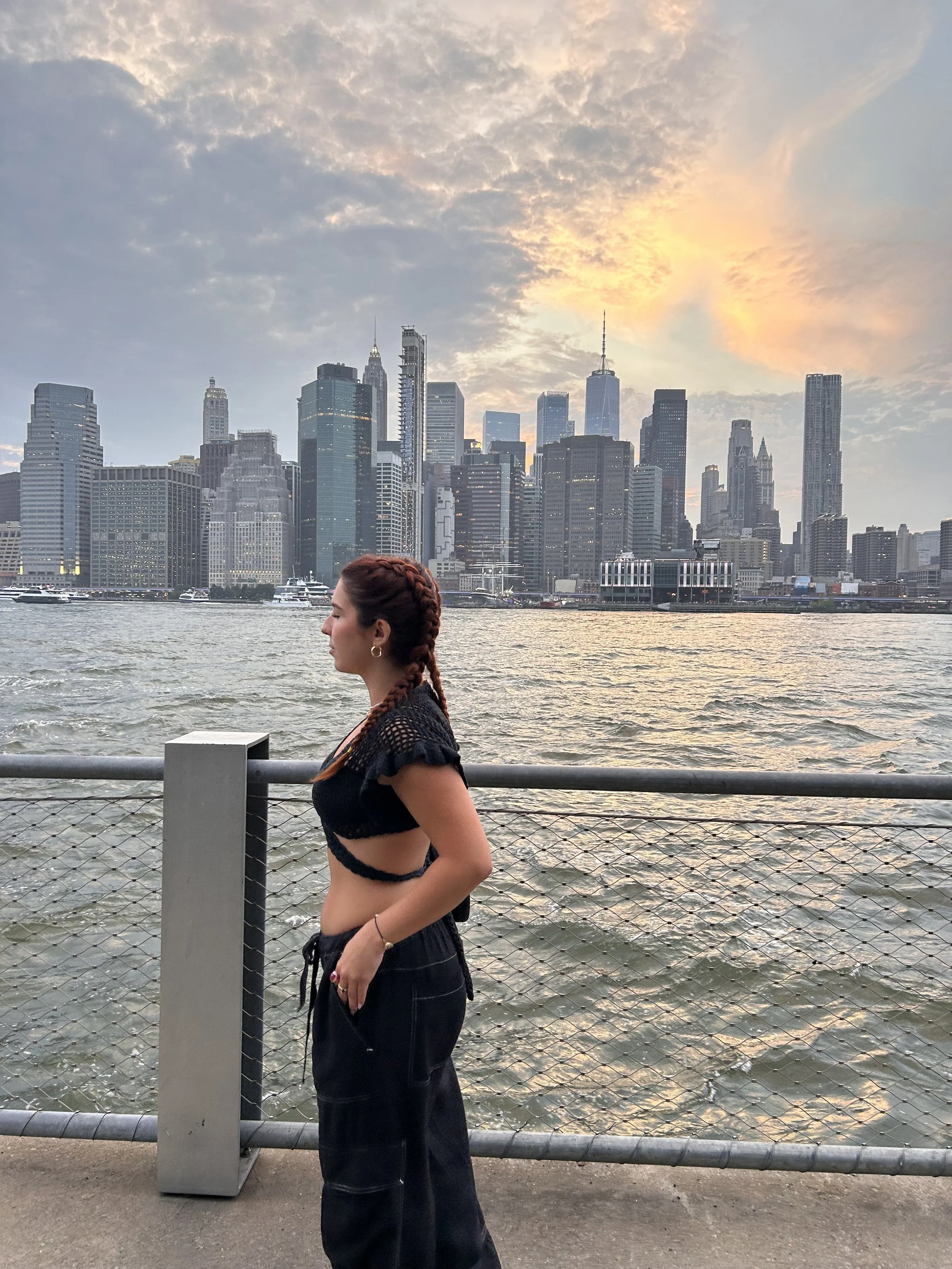 A woman with braided hair stands near a waterfront with a city skyline in the background, during sunset.