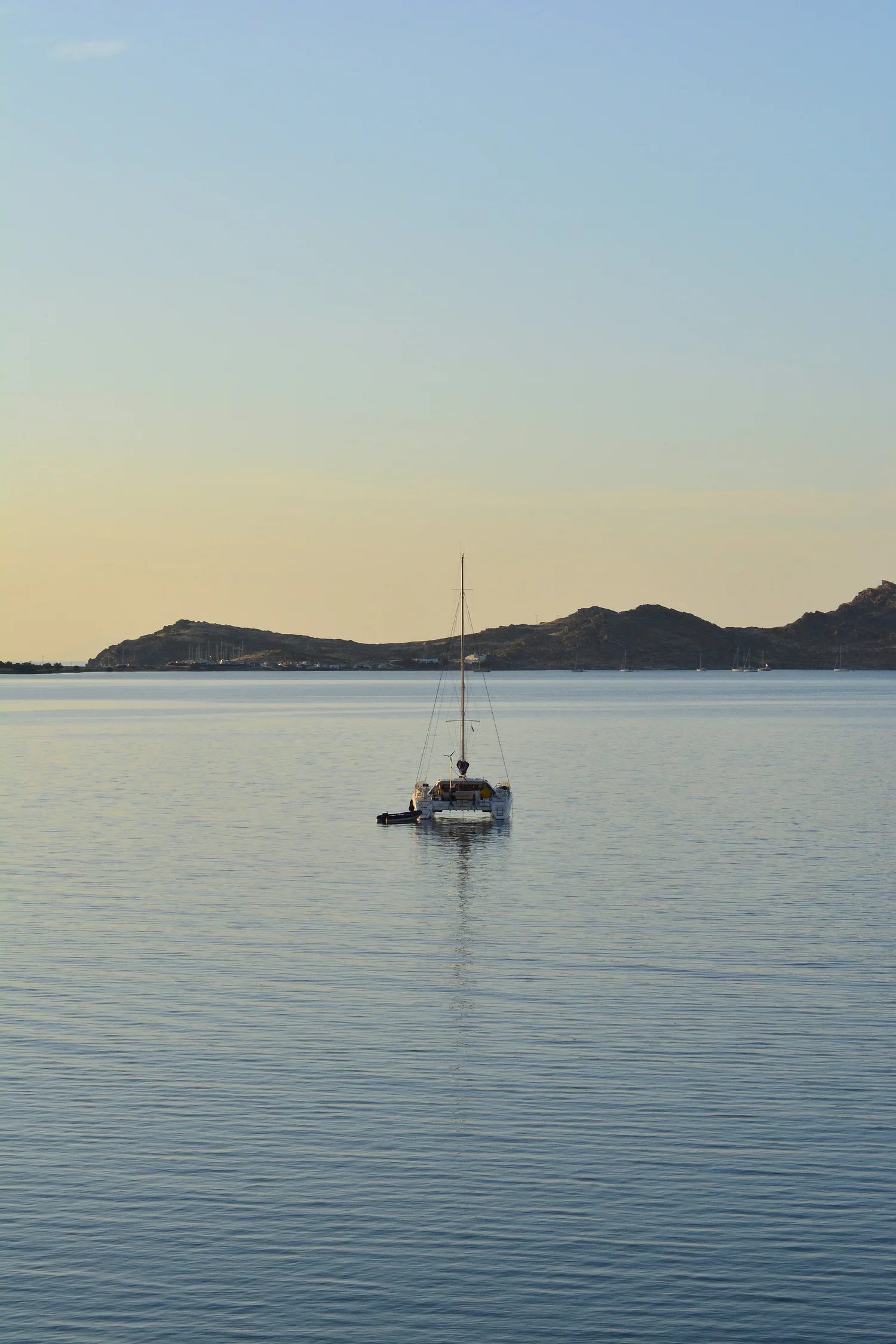 A sailboat floating on calm water near a shoreline with hills in the distance at dusk.