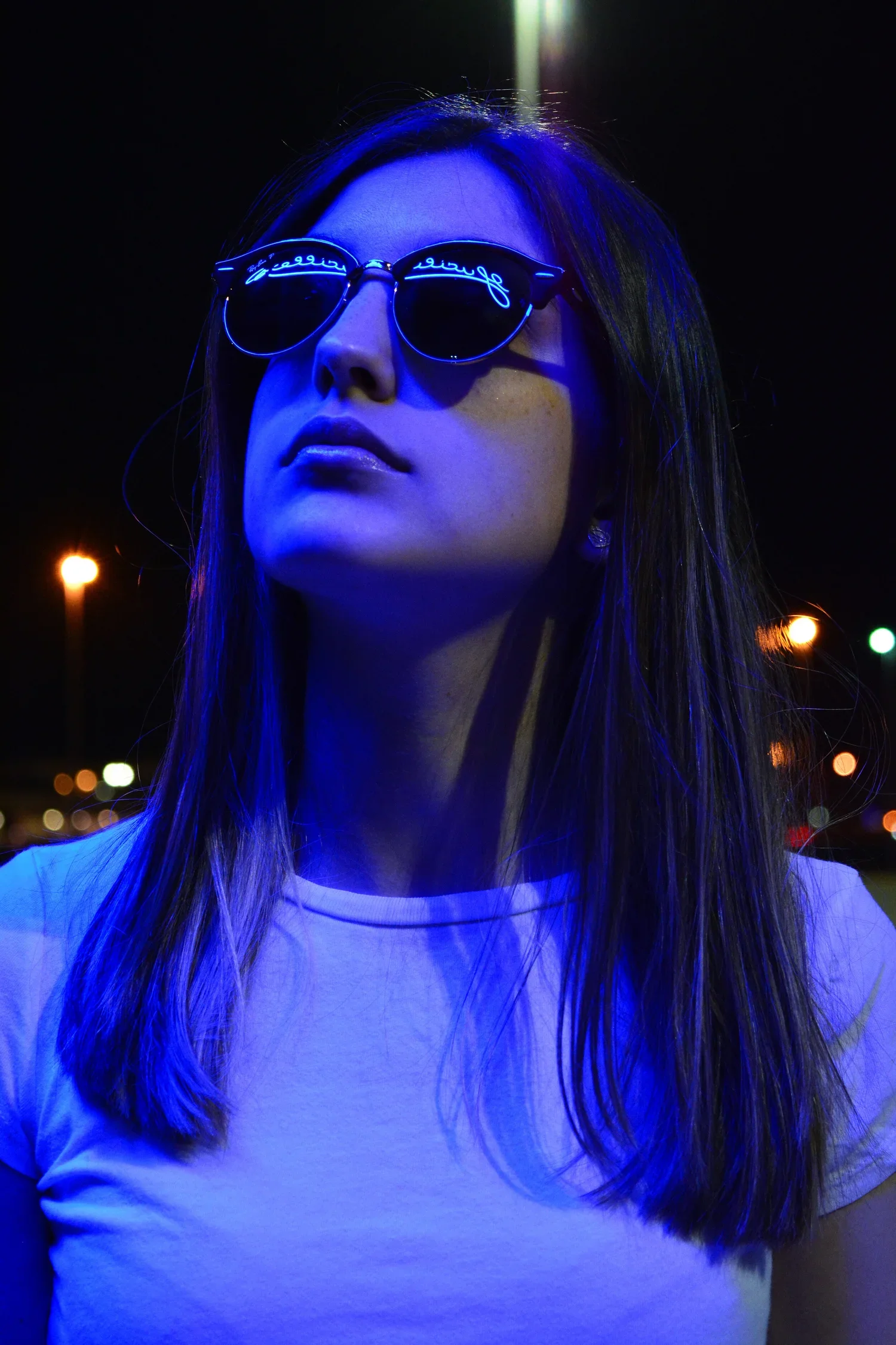 Nighttime portrait of a woman wearing sunglasses and a white t-shirt, illuminated by blue and yellow lights with blurred city lights in the background.