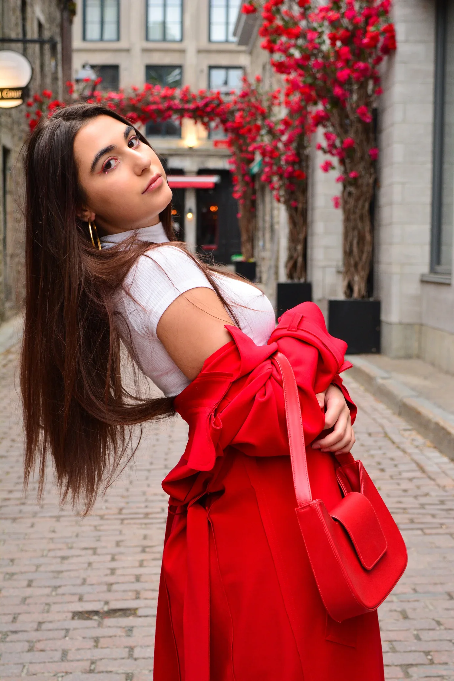 A woman with long brown hair wearing a white top with cut-out shoulders, gold hoop earrings, and bold eye makeup stands on a cobblestone street holding a red jacket and matching red purse. Pink flowering trees and building facades are in the backgrou