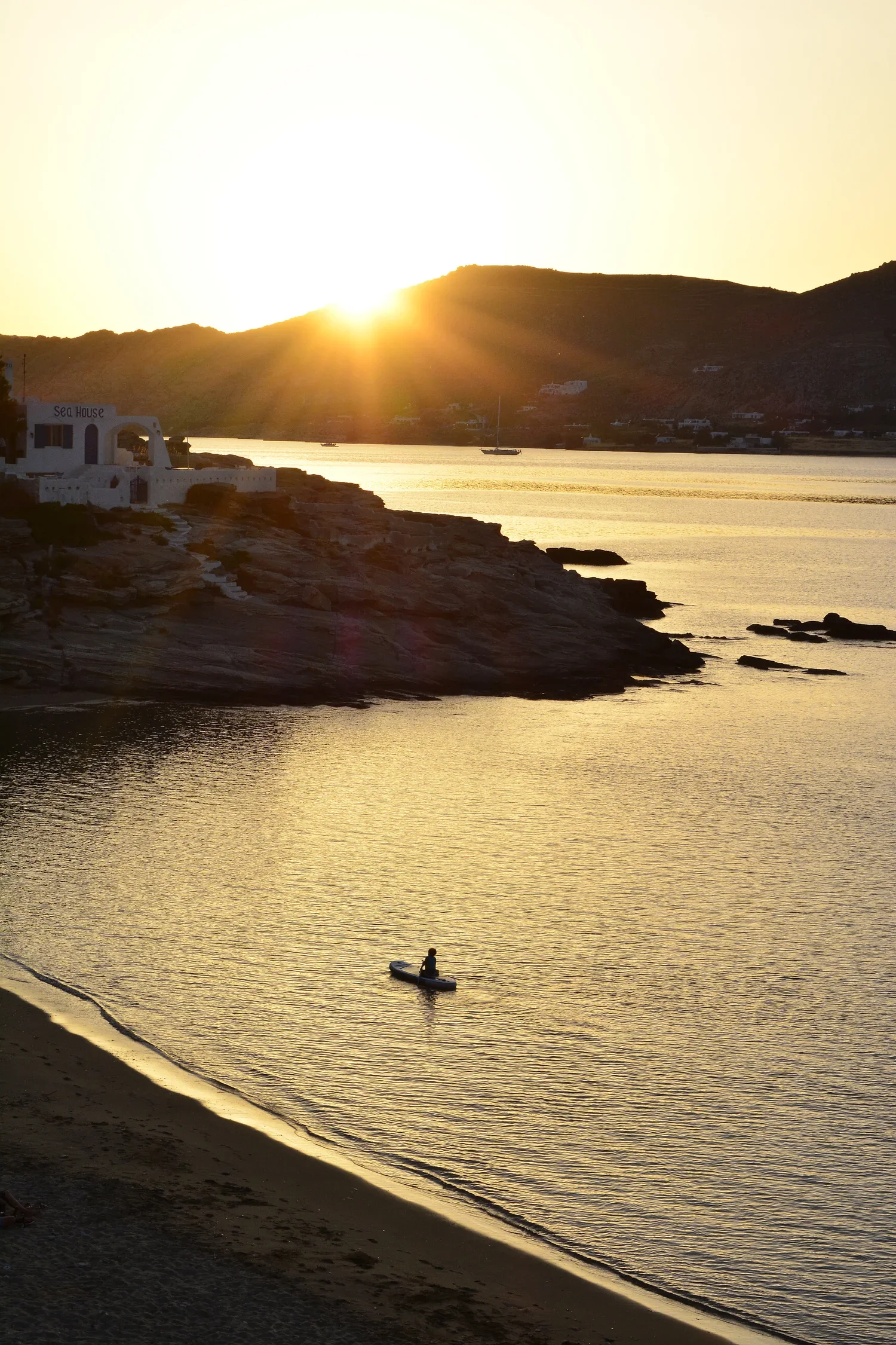 Sunset over a coastal landscape with a person paddleboarding on the water, a small beach in the foreground, rocky cliffs, and white buildings on the left side, with a sailboat in the distance.